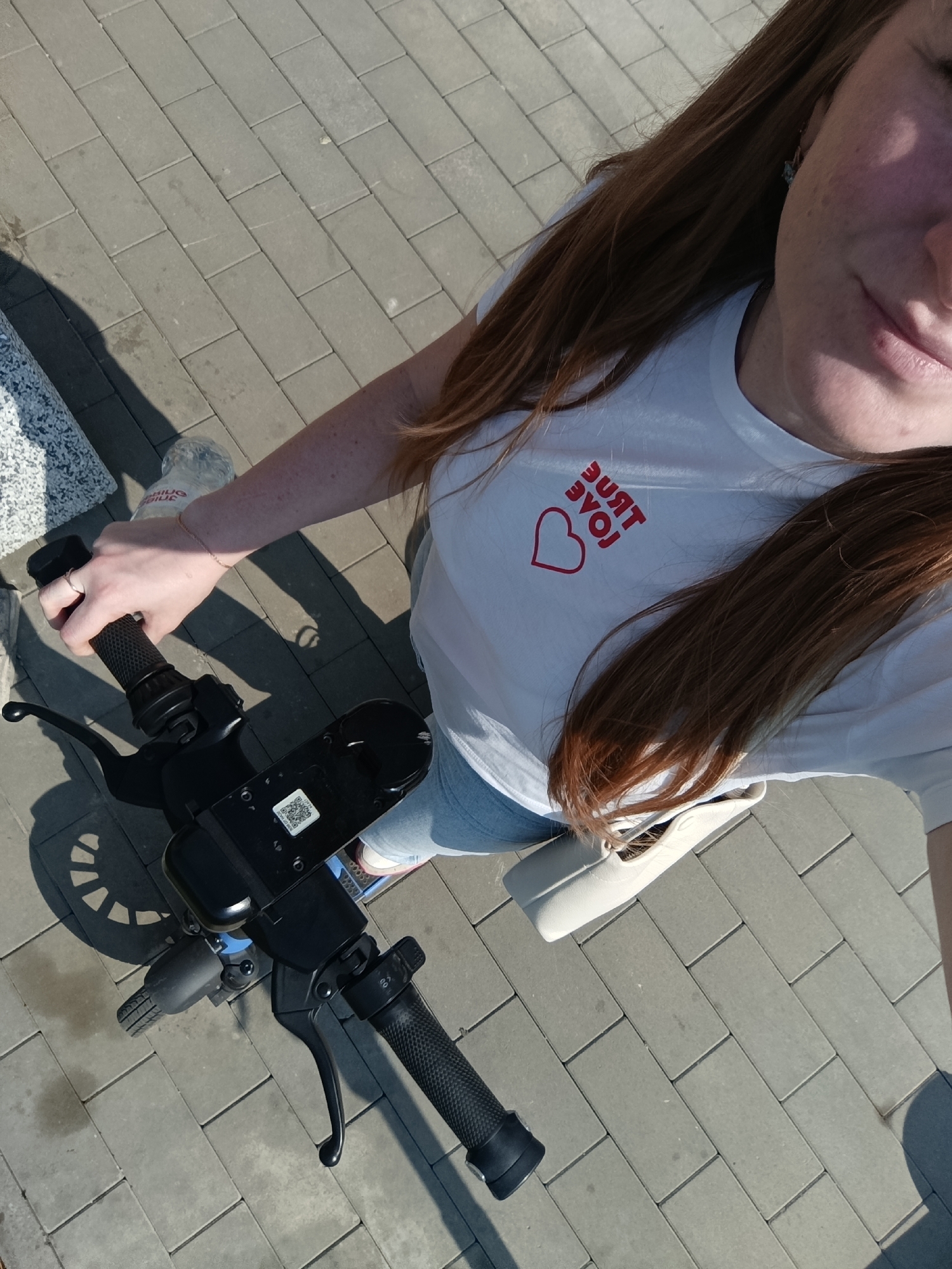 The image shows a woman, possibly taking a selfie, from an elevated perspective. She is standing on an electric scooter, holding onto the handlebars. She wears a white t-shirt with "TRUE LOVE" printed in red, along with a heart. She also wears jeans. A white purse is slung over her shoulder. The ground is paved with rectangular stones. A bottle of water is located near the scooter. A stone bench is visible in the upper left corner. The location is outdoors. The image contains a QR code on the dashboard of the scooter.