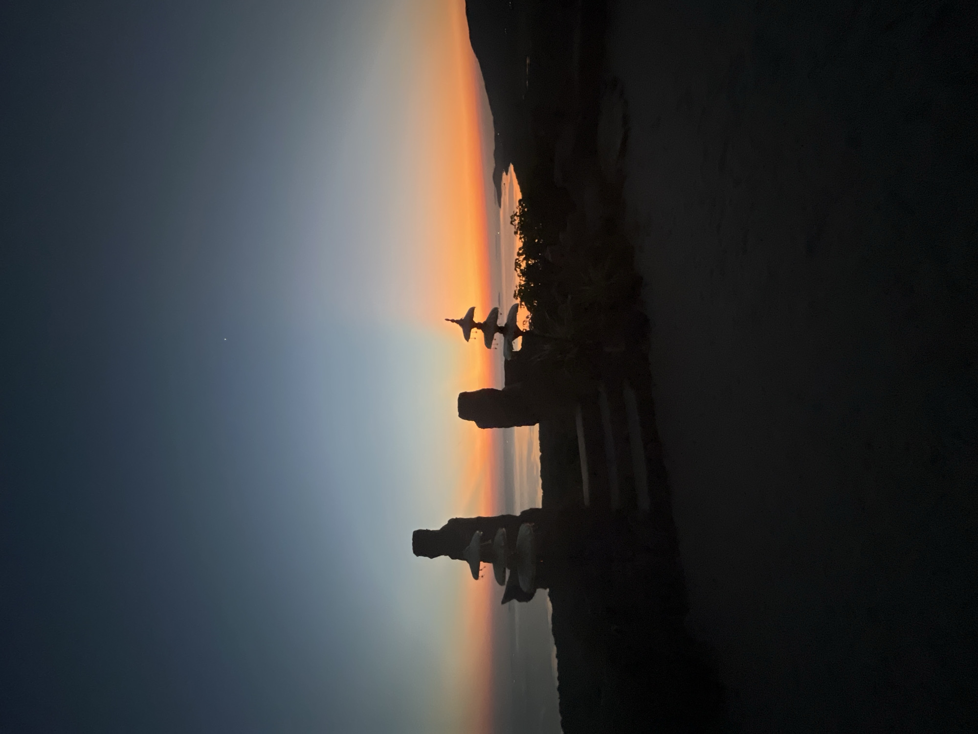 This is a vertical, low-angle, medium shot of a silhouetted landscape at dusk, likely taken from a high vantage point overlooking the ocean. In the foreground, two distinct stacked rock formations are visible, resembling abstract sculptures or cairns. The formation on the left is taller and more slender, with several flat, disc-like stones stacked atop each other. The formation on the right is shorter and wider, with a more irregular shape. Between these formations, a set of dark, stone steps descends into the foreground. The ground in the immediate foreground is dark and appears to be made of dirt or gravel.
The middle ground features a body of water, likely the ocean, reflecting the vibrant colors of the sunset. A distant coastline with rolling hills is visible on the right side of the frame. The sky transitions from a deep indigo blue at the top to a bright orange and yellow band just above the horizon, with subtle rays of light emanating from behind the distant hills. A single, faint star is visible in the upper left portion of the sky. Dense foliage is silhouetted against the bright horizon in the middle ground, surrounding the rock formations.
The scene appears to be located in the Karimunjawa Islands of Indonesia, a popular destination known for its natural beauty and coastal landscapes. The atmosphere is serene and tranquil, characteristic of a sunset in a tropical or coastal region. There are no people or animals visible in the image. The overall impression is one of natural beauty and a sense of peaceful solitude.