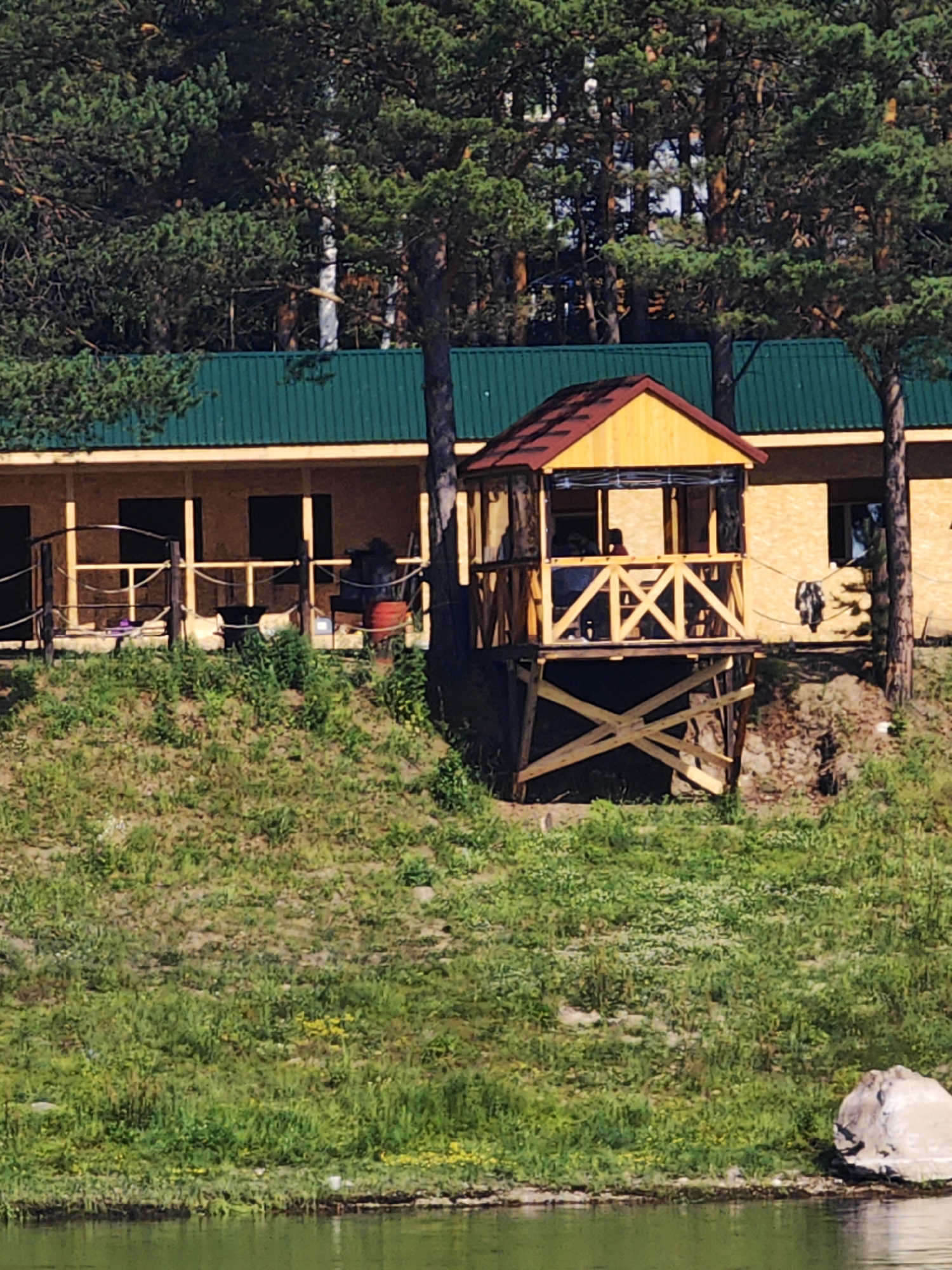 The image captures a serene natural setting with a wooden structure situated on a grassy slope overlooking a body of water. The structure, likely a house or a guesthouse, features a bright yellow facade and a green metal roof, with a prominent wooden gazebo perched on stilts on its left. The gazebo has a red-brown roof and open sides, offering a view of the surrounding landscape. The slope is covered in lush green grass dotted with yellow wildflowers. Pine trees form a dense backdrop to the building, suggesting a location in a forested area. In the foreground, the calm, green water reflects the sky and the trees. The overall impression is one of a peaceful rural retreat, possibly in Podporozh'e, Russia, known for its natural beauty and abundant forests.