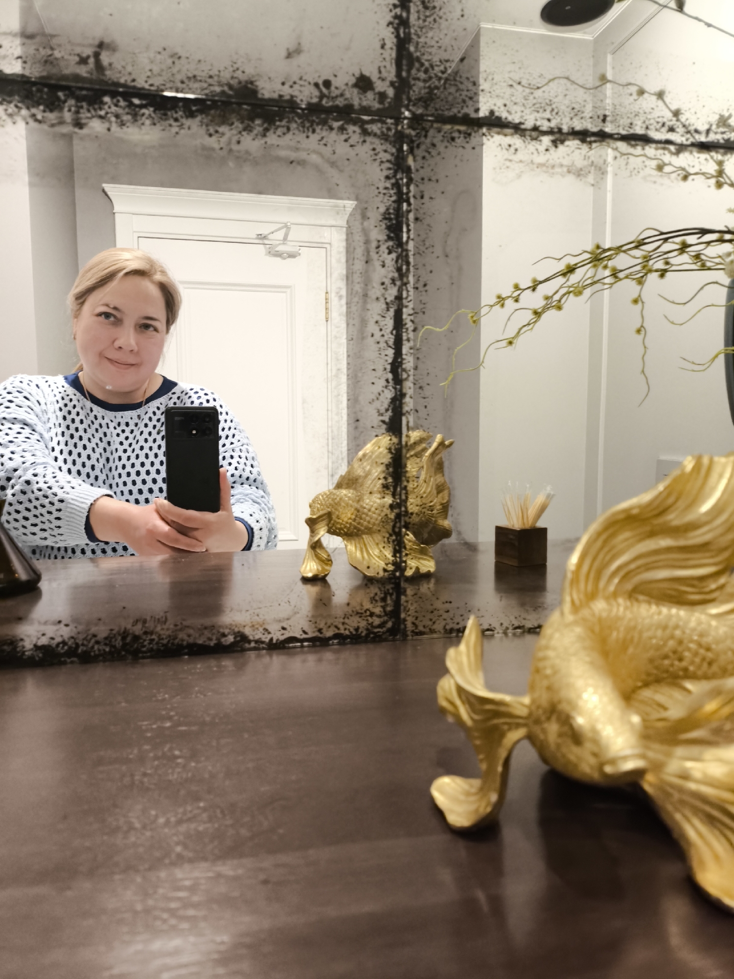 A woman is taking a selfie in a bathroom. She is holding a black smartphone in her right hand. The bathroom has a large, antiqued mirror with black markings. A door is visible in the background, as well as gold-colored decorative items, including fish sculptures. A small, dark wooden box with what appears to be cotton swabs sits on the counter. The countertop is a dark, brownish color. The woman is wearing a light blue, open-knit sweater with a dark blue neckline. The location is unknown.