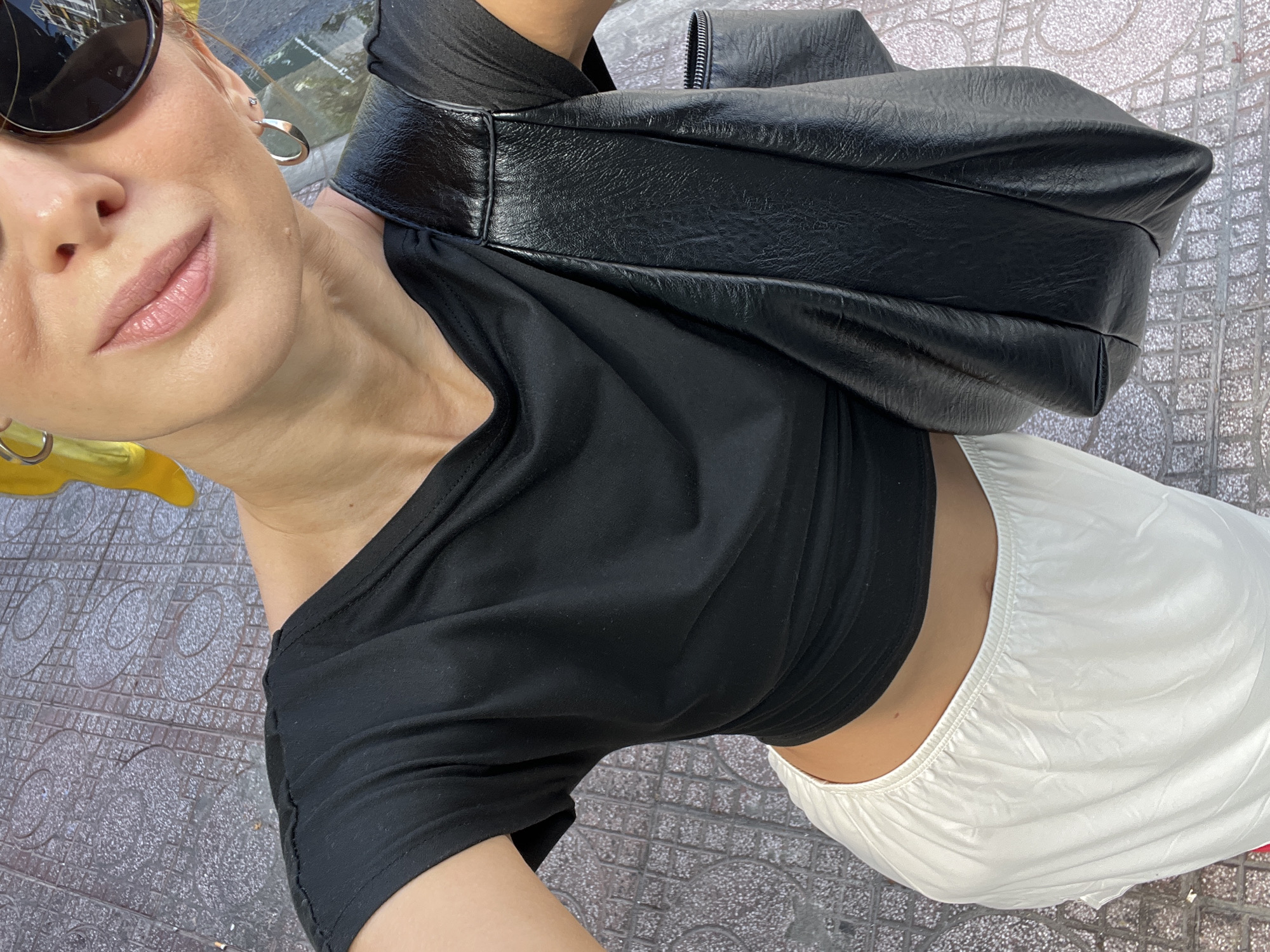 The image shows a close-up of a woman from the chest up, taken from a high angle outdoors. She is wearing a black t-shirt and a white skirt. A black leather handbag is slung over her right shoulder. She is also wearing sunglasses and a yellow hair accessory. The ground beneath her is paved with grey tiles. Based on the tiled pavement, the woman's attire and accessories, and the presence of outdoor elements, the scene appears to be a public space in the city of Nha Trang, Vietnam, possibly a street or walkway.