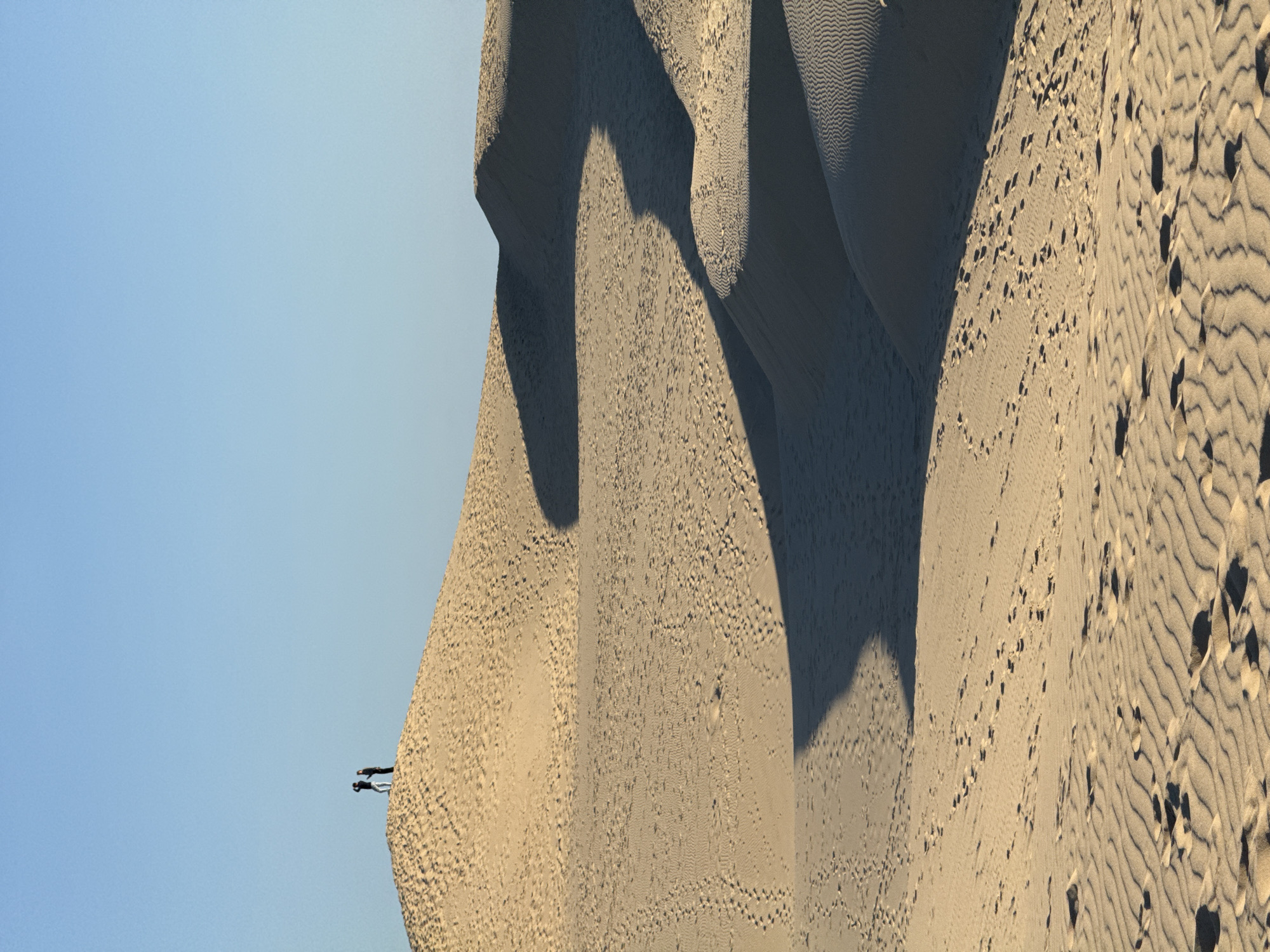 The image depicts the vast sand dunes of the Dune du Pilat in La Teste-de-Buch, France. The sun casts long shadows across the undulating sandy landscape, highlighting the rippled texture of the sand. Two individuals are visible standing on the crest of a dune in the upper left portion of the frame, silhouetted against a clear blue sky. The sandy terrain is marked with numerous footprints, suggesting recent human activity. The scene is serene and expansive, conveying a sense of scale and natural beauty.
