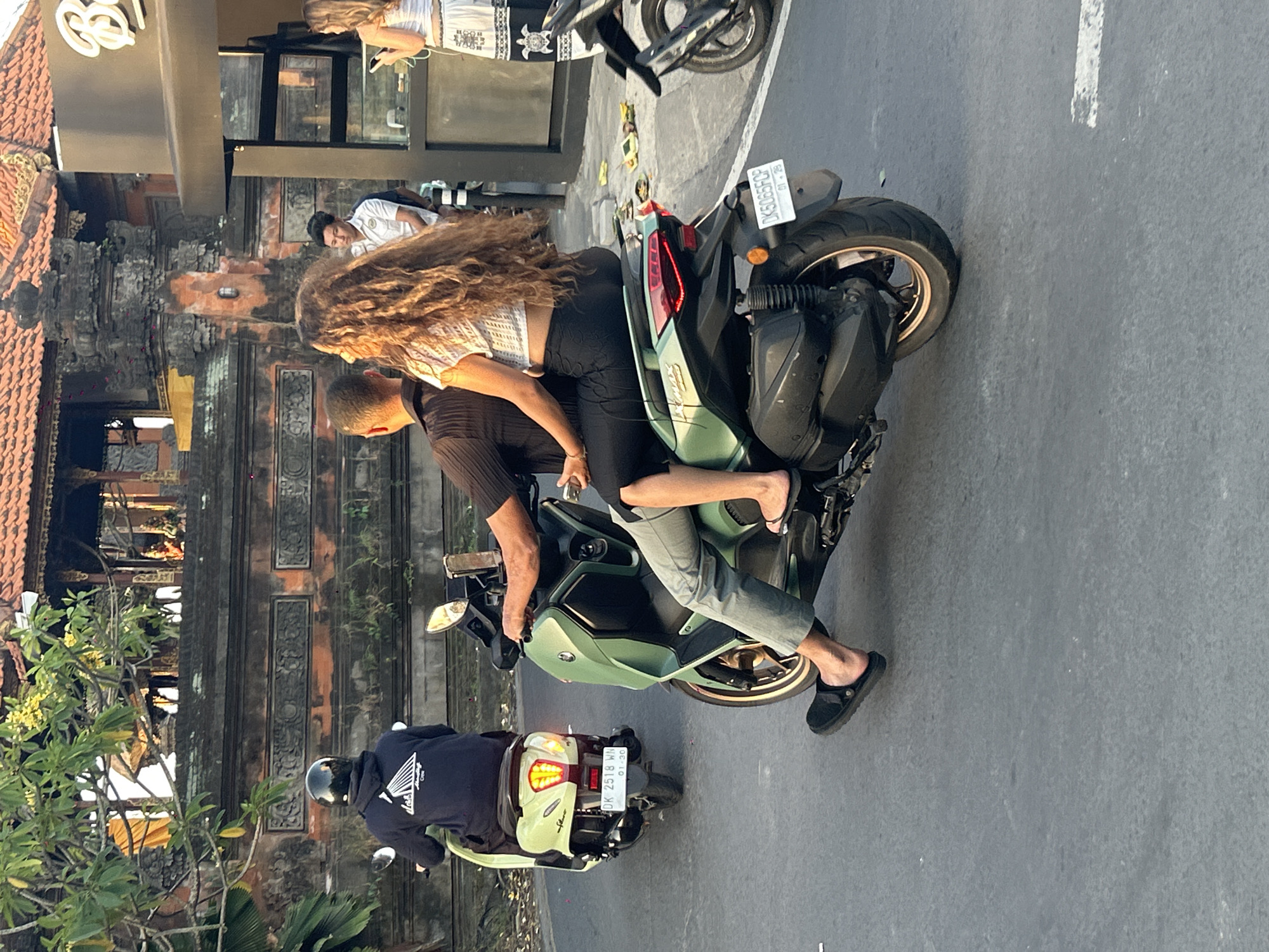 In North Kuta, Indonesia, at street level, a couple rides on a green scooter. The male rider, wearing a black shirt and grey pants, is in the foreground, with his left foot clad in a black clog resting on the ground. The female passenger, with long, curly brown hair, wears a white crop top and black pants, sitting behind him. A second scooter, a light green one, is visible to the left, carrying a person wearing a black hoodie and helmet. Behind the scooters, a building with a terracotta-tiled roof and intricate stonework is partially visible, with lush green trees and foliage surrounding it. The street is paved and appears to be an urban or semi-urban environment. A third scooter is parked or in motion to the right.