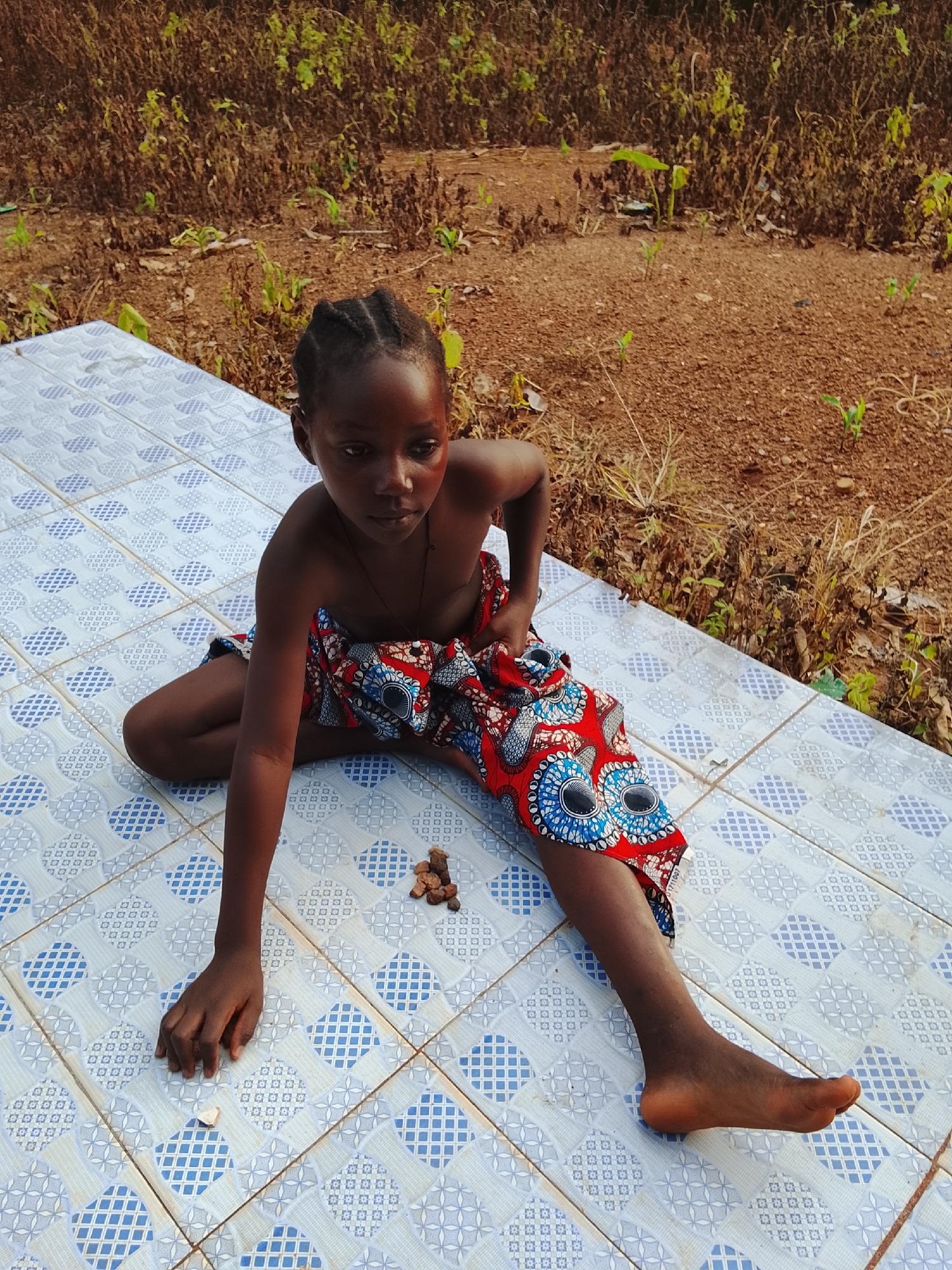 A young child, likely of African descent, sits on a tiled surface in a rural outdoor setting. The child is wearing a vibrant, patterned cloth around their waist, revealing their bare chest and arms. Their hair is styled in braided cornrows. In front of the child are several small, brown stones. The tiled surface is light-colored with a repeating blue diamond pattern. Behind the child, the ground is covered in dry soil and sparse vegetation, suggesting a dry or semi-arid environment. The background consists of dry grasses and plants, with a visible brown earth floor. The image appears to have been taken during daylight. The overall impression is one of a simple, rustic environment.