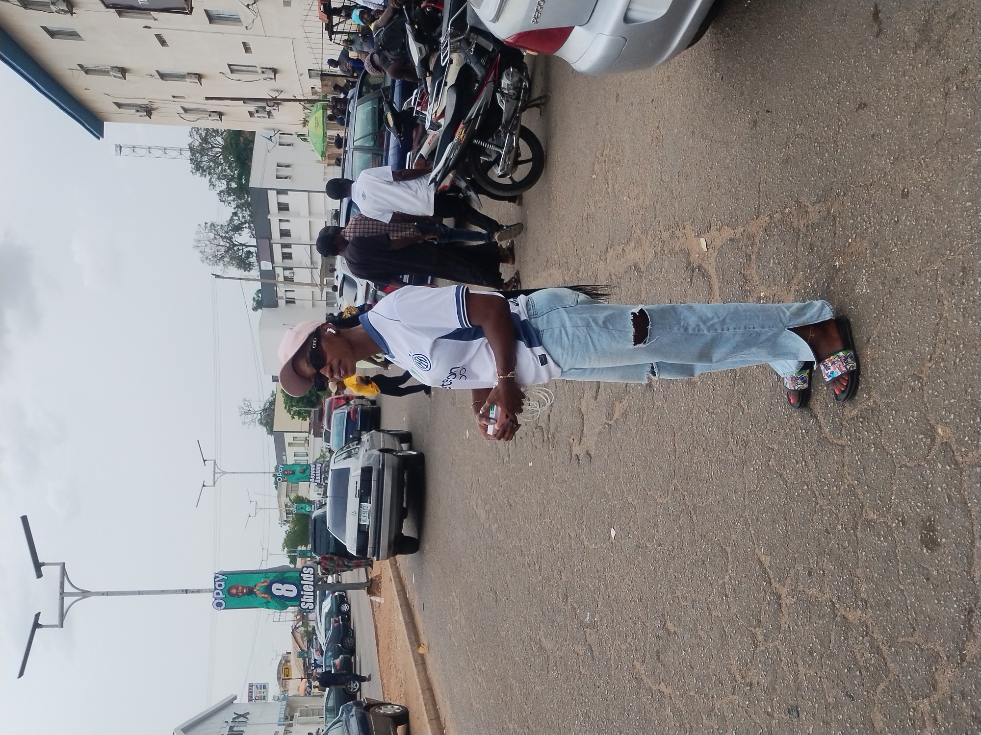A person stands on a street in Jos, Nigeria. They wear a white T-shirt, light blue ripped jeans, and patterned sandals. A pink hat and sunglasses are also visible. The street has several cars and motorcycles parked along the side. A building is visible in the background, along with light poles bearing "OPay" banners. Other people are present in the background. The street is asphalt and shows signs of wear.