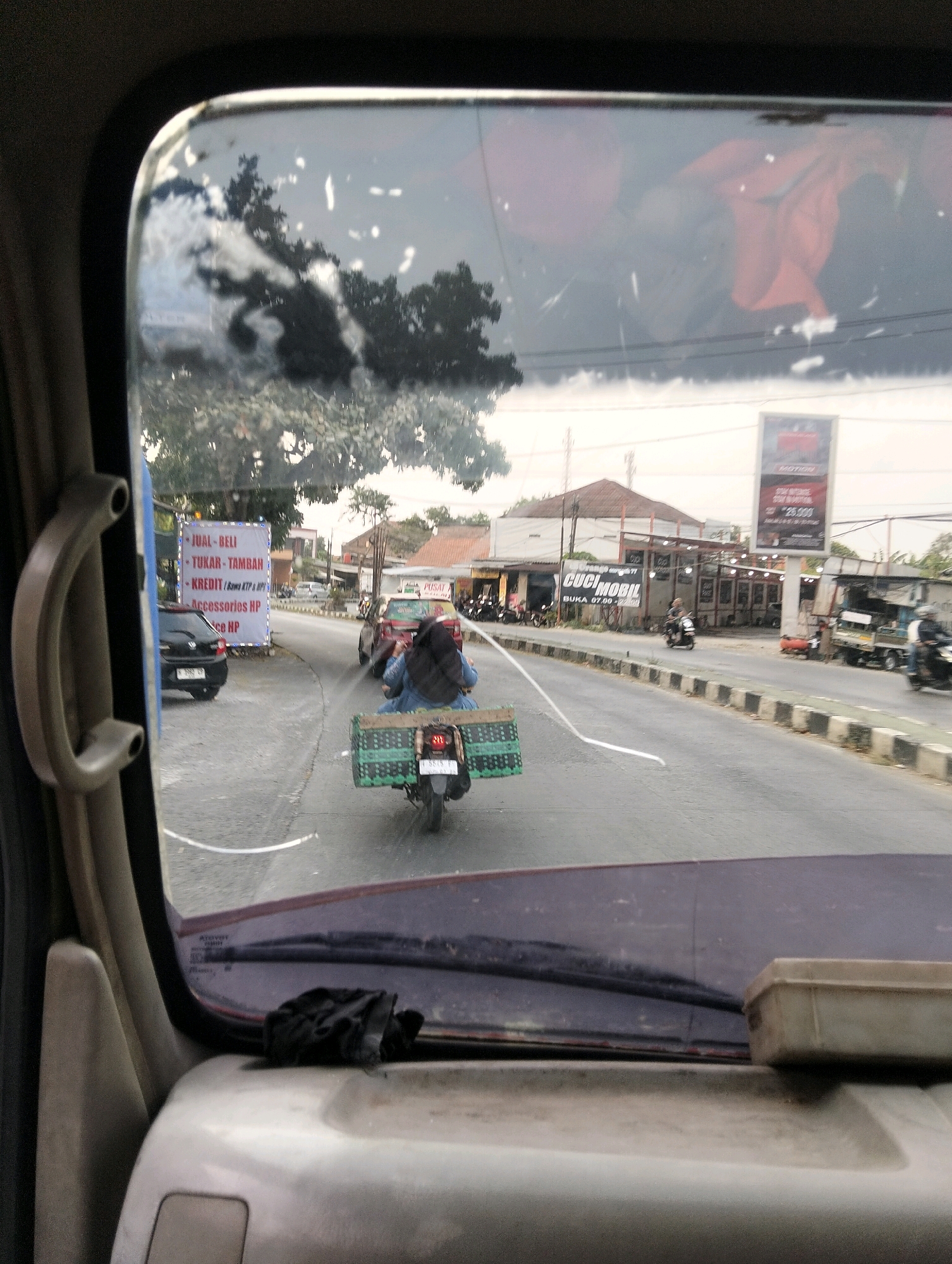 The image is taken from inside a vehicle, looking out through the windshield. The view shows a street scene in Semarang, Indonesia. On the left side of the street, there is a large white banner with text in Indonesian that appears to be an advertisement for "JUAL-BELI", "TUKAR-TAMBAH", and "KREDIT" services, possibly for mobile phones and accessories. Further down the road, there are cars and a scooter carrying a person with a large green basket. Across the street, there are buildings, including one with a sign that says "CUCI MOBIL" (Car Wash) and a large billboard. The overall impression is a busy street in an urban area.