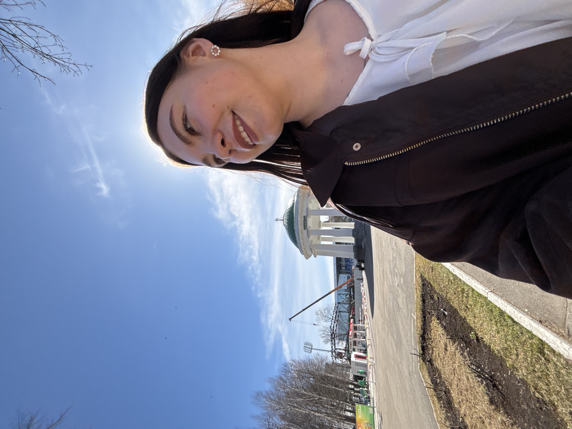 A woman with dark hair, a white blouse, and a dark jacket is smiling in the foreground. In the background is a blue sky with some clouds, a gray road, a green patch of grass, bare trees, a crane, and the rotunda in Perm's Gorky Park, Russia.