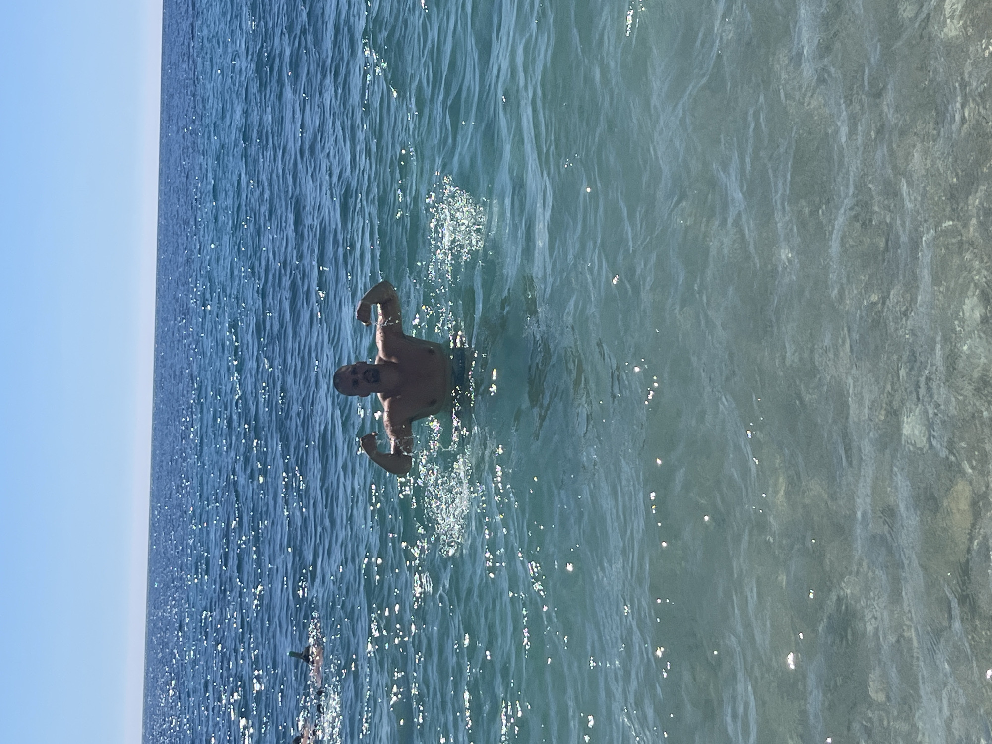 A man swims in the sea off the coast of El Poble Nou de Benitatxell/Benitachell, Spain. The water is clear and blue, reflecting sunlight. The sky is also blue and clear. The man is positioned in the middle of the frame. The water near the shoreline on the right is noticeably shallower.