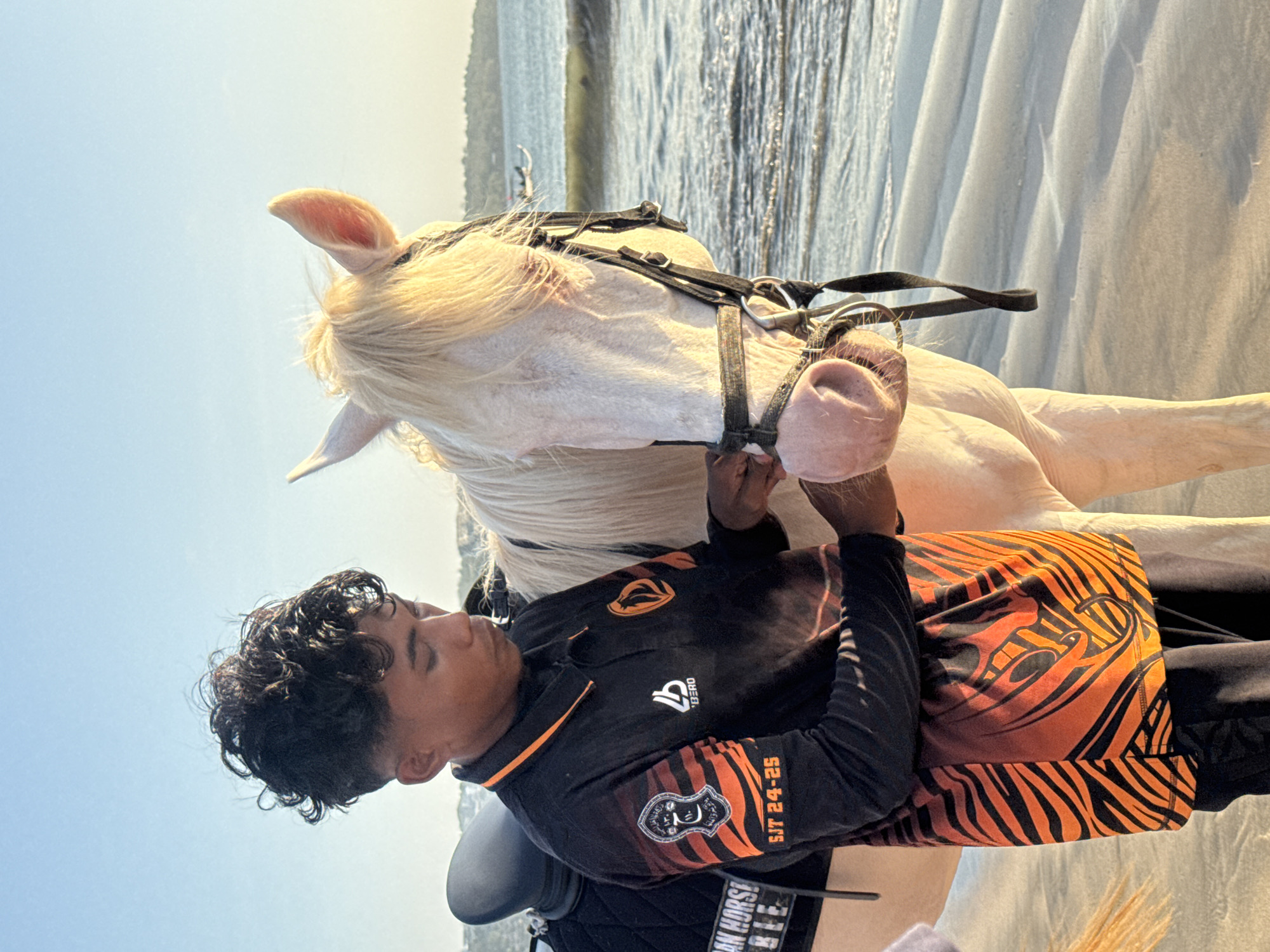 In Kuta Selatan, Indonesia, a young man with dark, curly hair, wearing a black polo shirt with orange tiger stripe accents and an emblem on the left sleeve, stands beside a light-colored horse on a sandy beach. He is looking down at the horse, appearing to adjust its bridle. The horse, with a light mane and pink muzzle, is facing the camera. A saddle is visible on the horse's back. The background shows the ocean with gentle waves and a distant shoreline with vegetation and structures. The sky is clear and blue, illuminated by the warm light of what appears to be a sunset. The overall scene depicts a moment of interaction between the man and the horse by the sea.