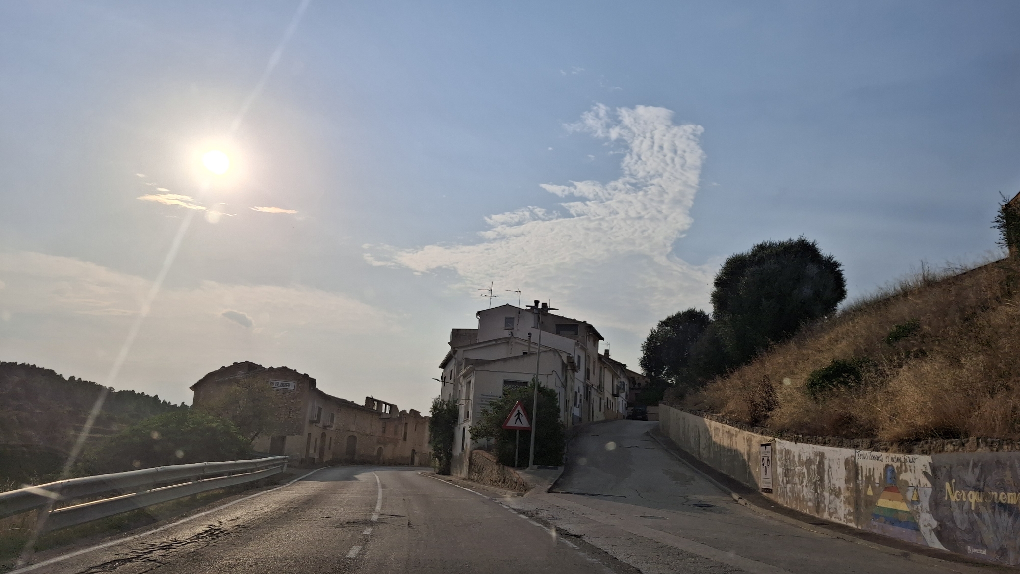 The image captures a rural road in Valjunquera, Spain, under a bright, sunny sky. The road curves to the left, with a metal guardrail on the left side and a dry, grassy hillside on the right. Several buildings are visible, appearing to be older, stone structures. On the left, a rustic building with a weathered facade and a sign is prominent. Further down the road, on the right, a cluster of white buildings forms the center of a small town or village. One of these buildings has a pedestrian crossing sign in front of it. The hillside on the right is adorned with a mural on a low concrete wall, featuring various symbols and text, including what appears to be a rainbow pyramid and the words "Tots tenim el mateix futur" and "Norquremrem". There are no people or animals clearly visible in the scene. The overall impression is one of a quiet, sun-drenched day in a Spanish rural setting.