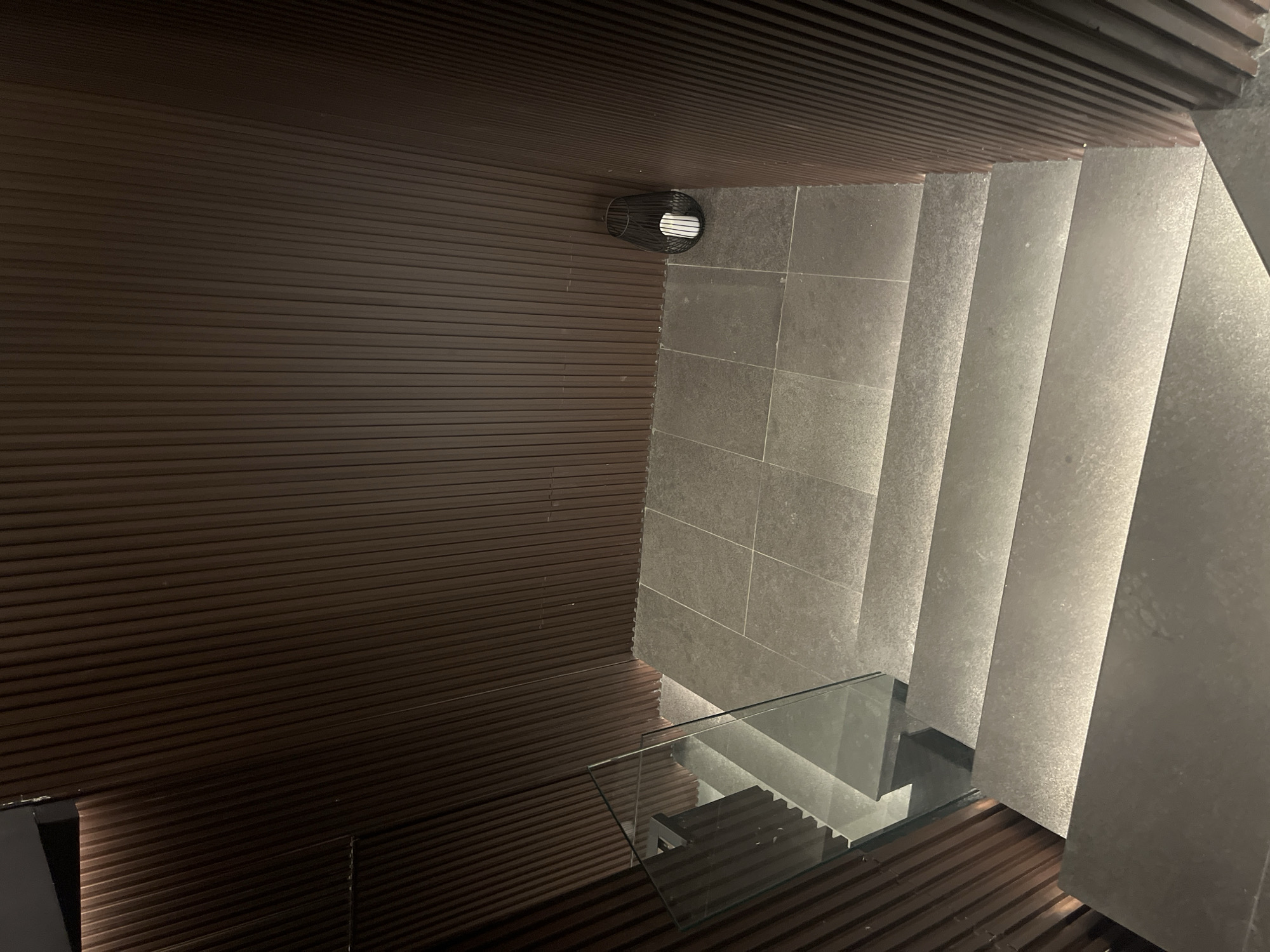 Captured from an elevated perspective, the image showcases an interior architectural design. The left side features a dark brown wall constructed from horizontal slats. On this wall, a cylindrical wireframe light fixture is mounted, emitting a soft glow. Adjacent to the slatted wall, a grey stone staircase ascends, lit subtly. A glass railing provides a modern touch to the staircase. The floor is also constructed from the same horizontal slats as the wall, suggesting a cohesive design. There is no evidence of any specific people, subjects, or actions occurring in the image. The location is unknown.