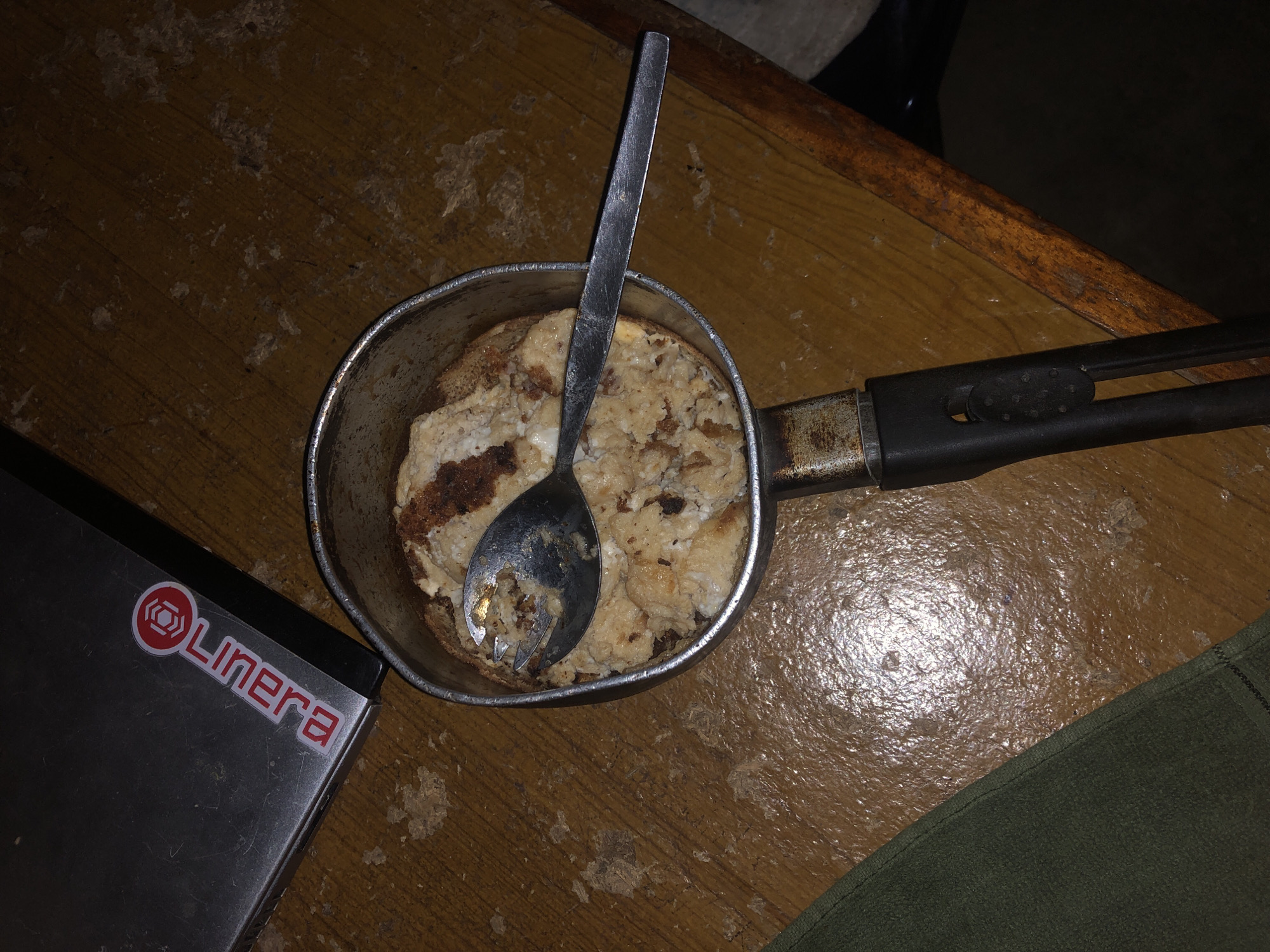 An overhead shot shows a small, stainless steel pot with a black handle containing a serving of what appears to be oatmeal or porridge. A silver spoon is resting in the pot, partially submerged in the food. The pot is positioned on a textured, wooden surface, possibly a table, which has a worn and stained appearance. To the upper left of the pot, a dark grey, flat object with a red and white logo is visible. A green cloth is draped on the lower left side of the frame. The lighting is dim, casting shadows and highlighting the textures of the surfaces. The scene is devoid of any people or discernible urban elements.
