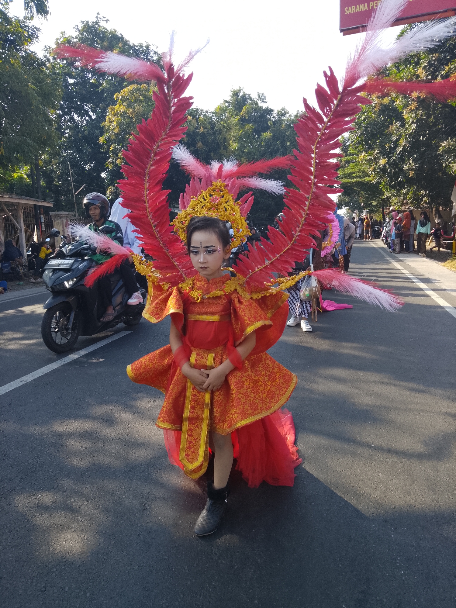 This is a medium shot, outdoor, eye-level photograph taken on a paved road, possibly in Buduan, Indonesia, during daylight hours. The foreground is dominated by a young child in an elaborate costume. The child, who appears to be female, is wearing a bright red and gold costume with large, decorative feather-like wings extending from the back. The costume features a golden headdress and intricate gold trim. The child has dark hair and is wearing dark makeup on her eyes and eyebrows. She is looking directly at the camera with a serious expression and has her hands clasped in front of her. She is wearing black boots.
To the left of the child, a dark-colored motorcycle is visible, with a person wearing a helmet and a green shirt riding it. Behind the motorcycle, other people and a fence are partially visible.
The background of the image shows a road with a white line marking the lane. On the right side of the road, a line of people are standing, facing towards the left, observing something. Some of them are dressed in casual clothing, while others appear to be wearing traditional attire. Trees with green foliage line the right side of the road, and a blue sky with some white clouds is visible above. A red sign with white lettering, partially obscured, is present in the upper background, with the visible text "SARANA PER". The overall scene suggests a parade or cultural event.