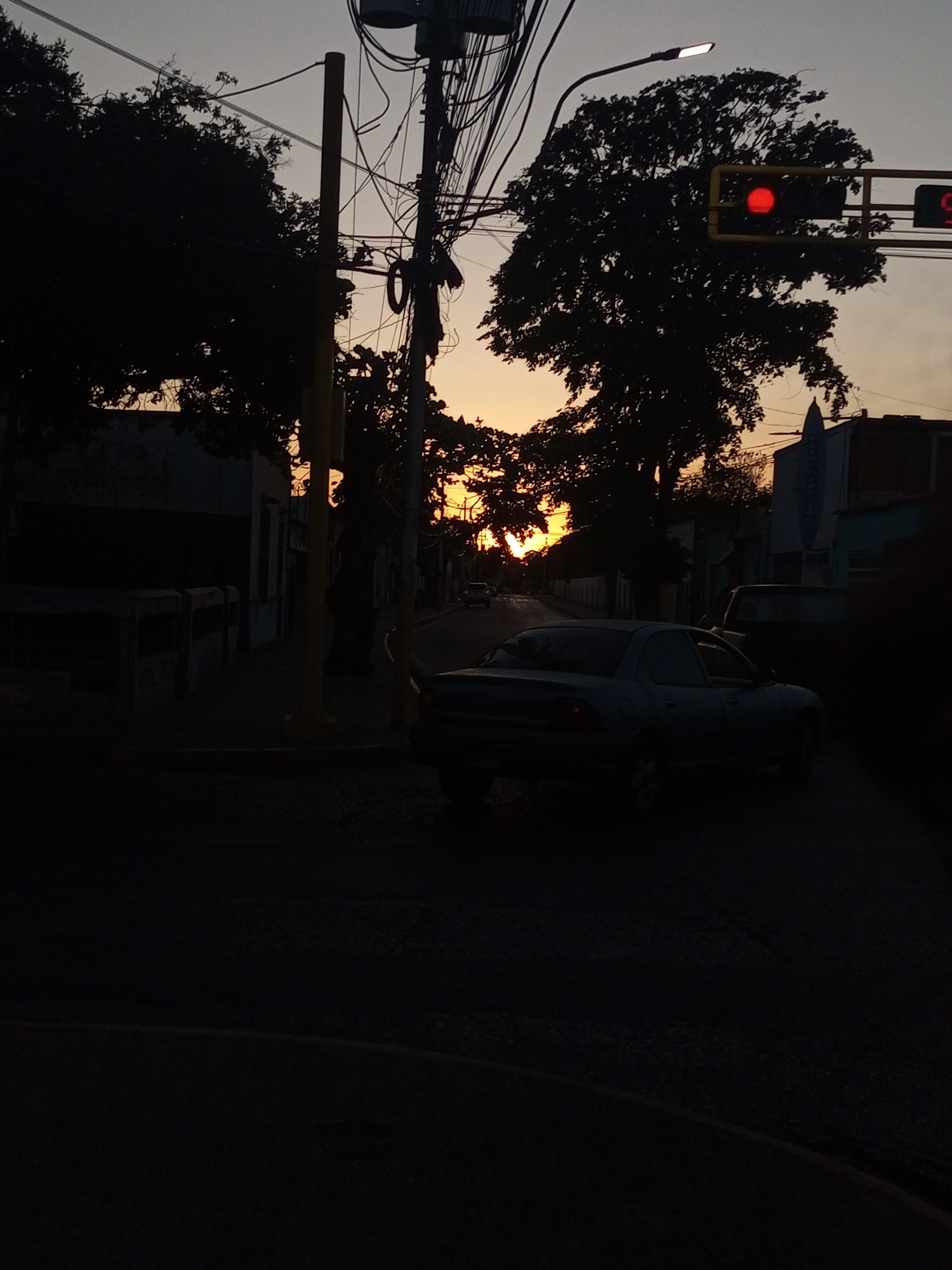 The image depicts a street in Juan Griego, Venezuela, at dusk. A car is parked on the right side of the street, with other vehicles visible further down. Traffic lights are overhead, showing red. Silhouetted trees frame the view, with the setting sun visible through the branches. Buildings line the street, and power lines crisscross above. The overall scene is dark, with details obscured by shadows.