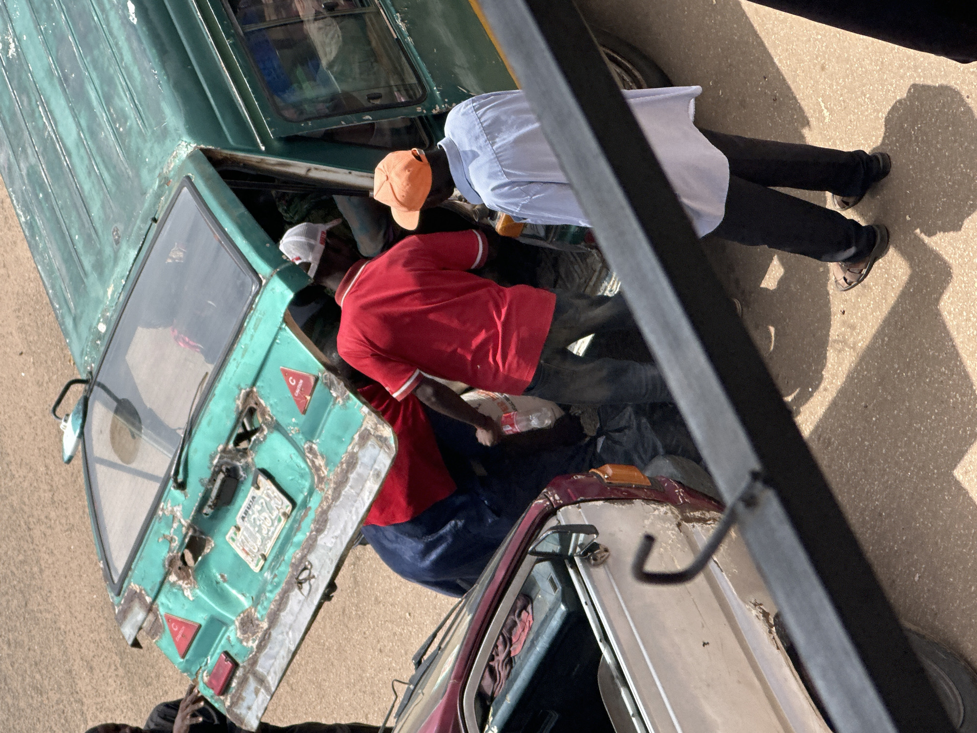 The image shows a group of people gathered around a damaged green minibus and a smaller, silver and maroon vehicle in what appears to be a street in Bukuru, Nigeria. The rear hatch of the green minibus is open and heavily rusted, revealing signs of damage. A license plate reading "ABUJA HUJ 3528C" is affixed to the hatch. Three individuals are visible: one wearing a red shirt and white cap, another in a light blue shirt and orange cap, and a third partially obscured person in a dark shirt. The red-shirted individual is seen interacting with the interior of the minibus. The silver and maroon vehicle to the left is also damaged, with its doors open and its interior exposed. The ground is paved with asphalt or concrete, and sunlight casts shadows, indicating an outdoor daytime scene.