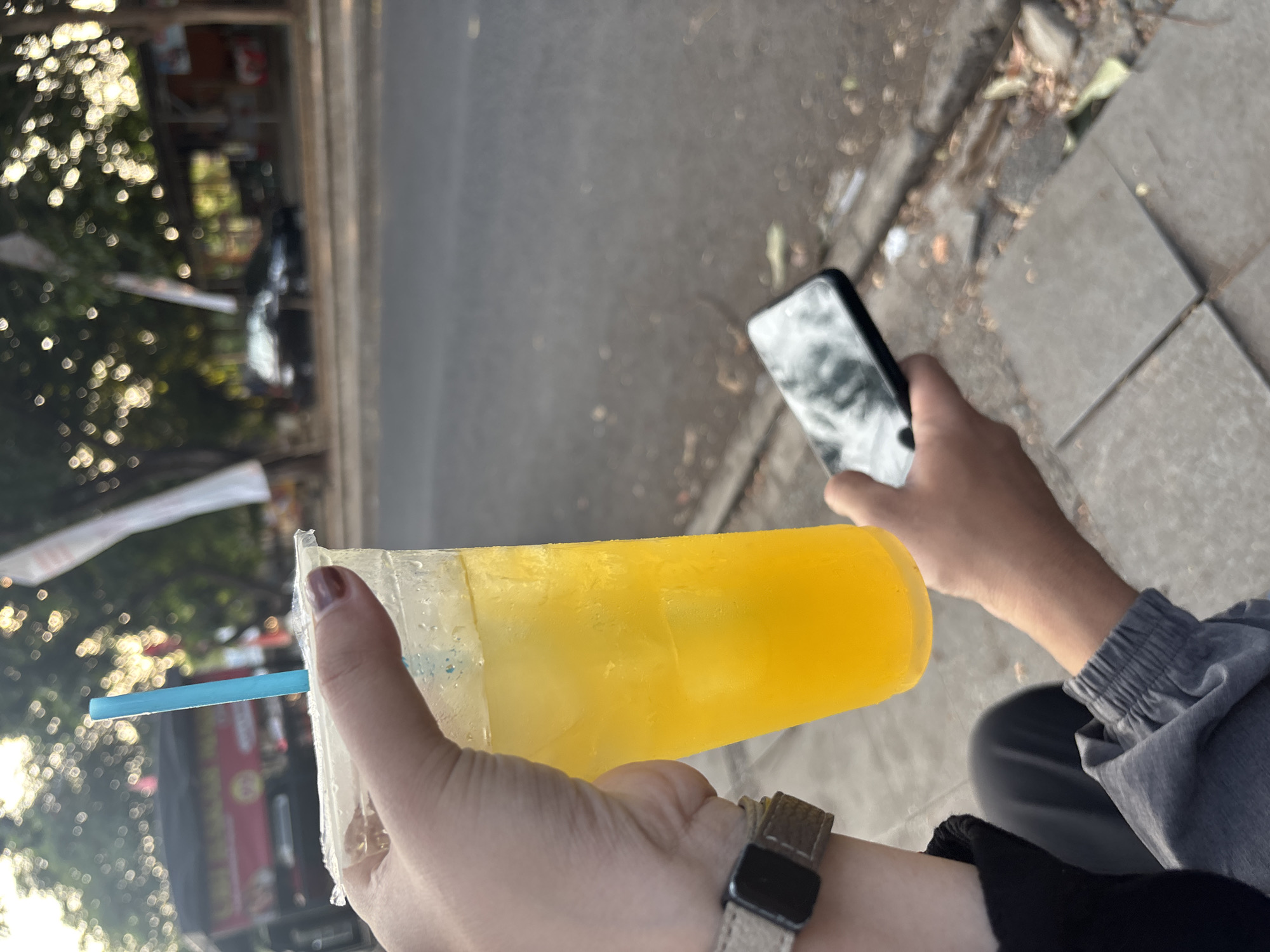In Cirebon, Indonesia, a person's hand holds a clear plastic cup filled with a bright yellow beverage and ice, with a blue straw inserted. The person is also holding a dark-colored smartphone in their other hand. They are seated on a paved surface, likely a sidewalk, with a street and blurred background of trees and structures. The lighting suggests it is daytime, possibly late afternoon, given the warm glow.