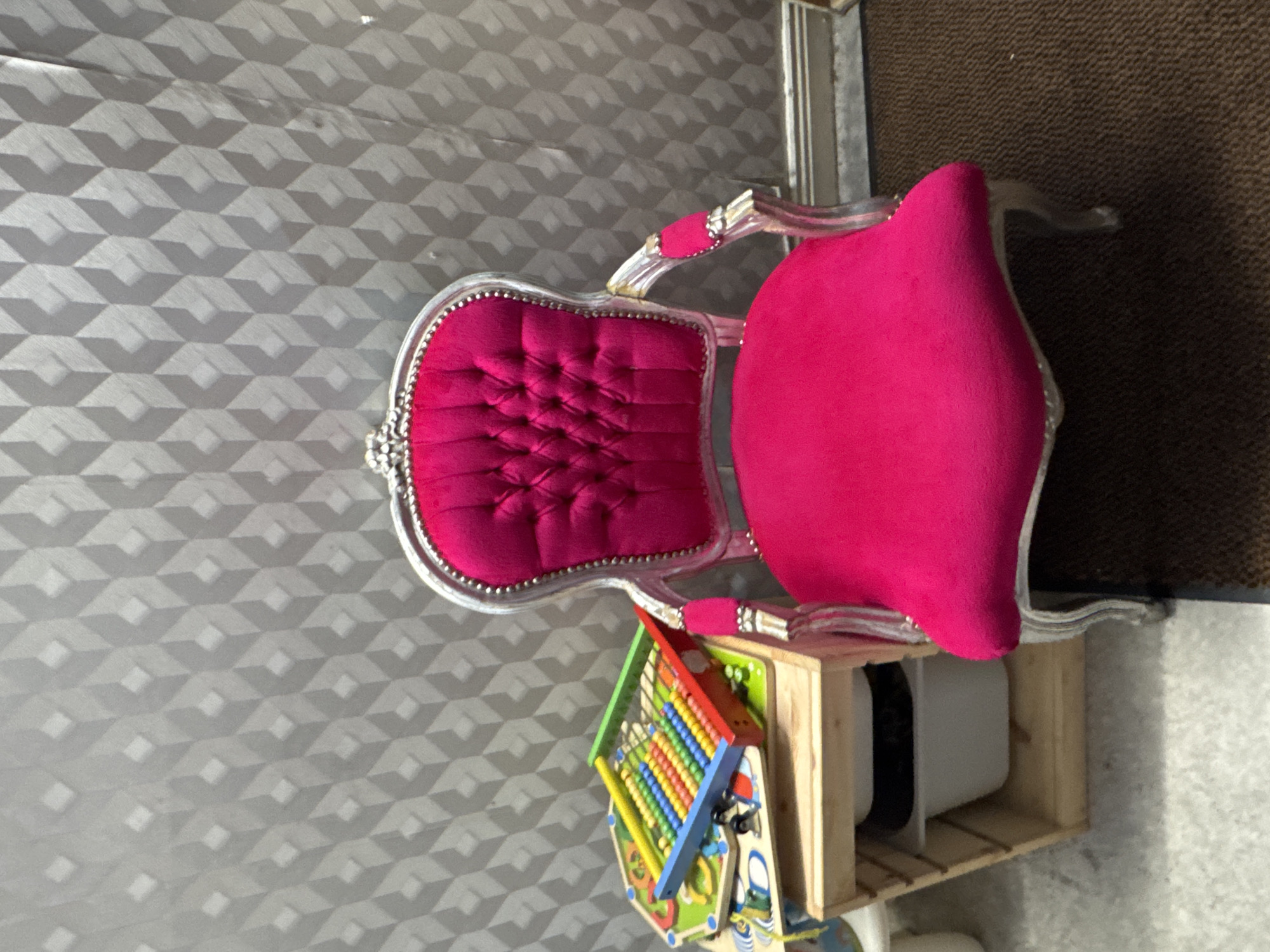 A pink tufted armchair with a silver ornate frame sits next to a wooden shelf unit. On the shelf unit, a colorful abacus and other children's toys are placed. The background is a grey textured wall with a geometric pattern. The floor is carpeted in brown. The scene appears to be indoors, likely in a child's play area or a classroom. This image is not specific to Clonmel, Ireland.