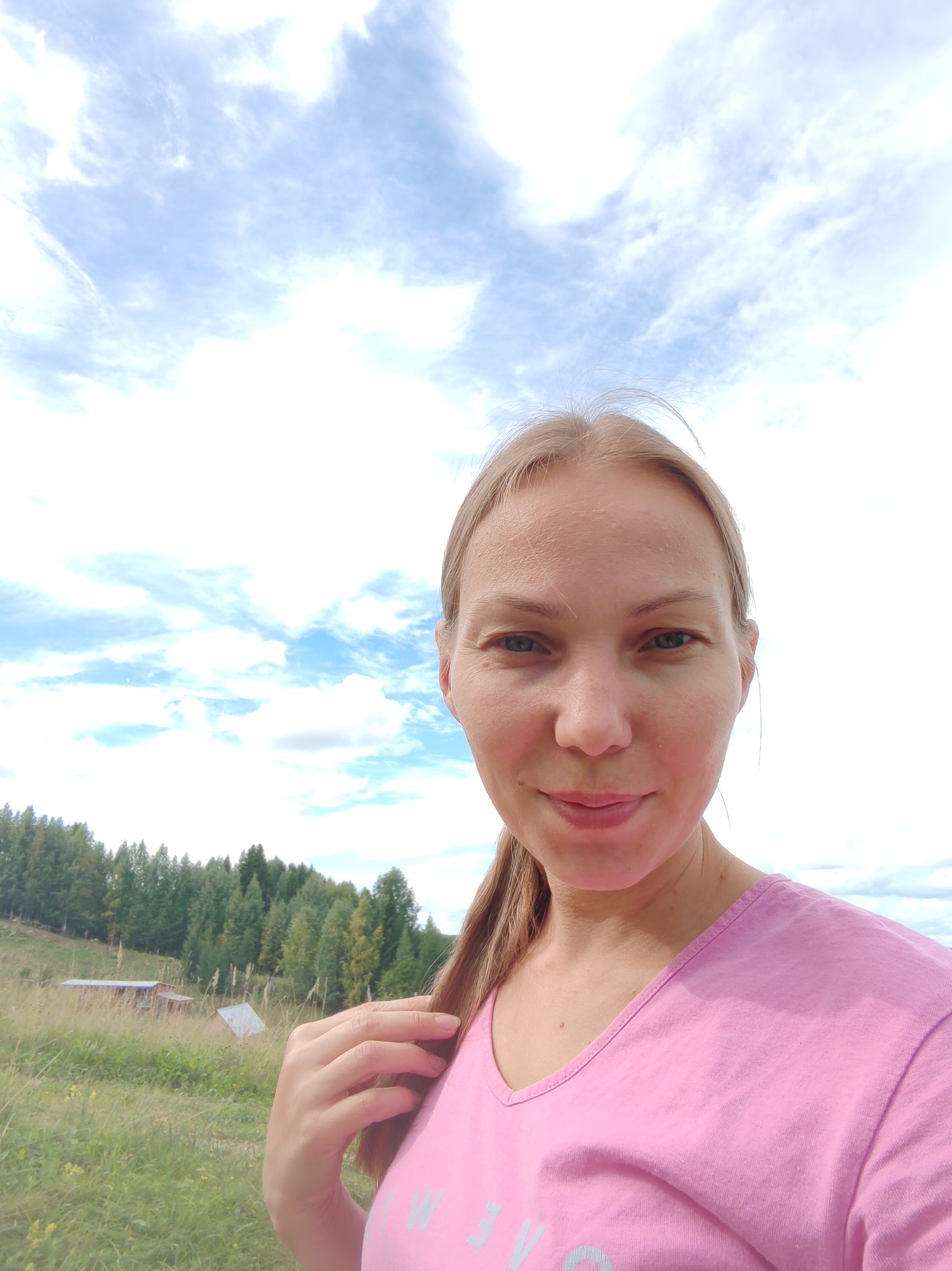 A woman with blonde hair and blue eyes, wearing a pink t-shirt, takes a selfie outdoors. She is standing in a grassy field with trees in the background. The sky above is partly cloudy. Two small buildings, possibly sheds, are visible in the middle ground to the left of the frame. The location is identified as Chukhlem, Russia.
