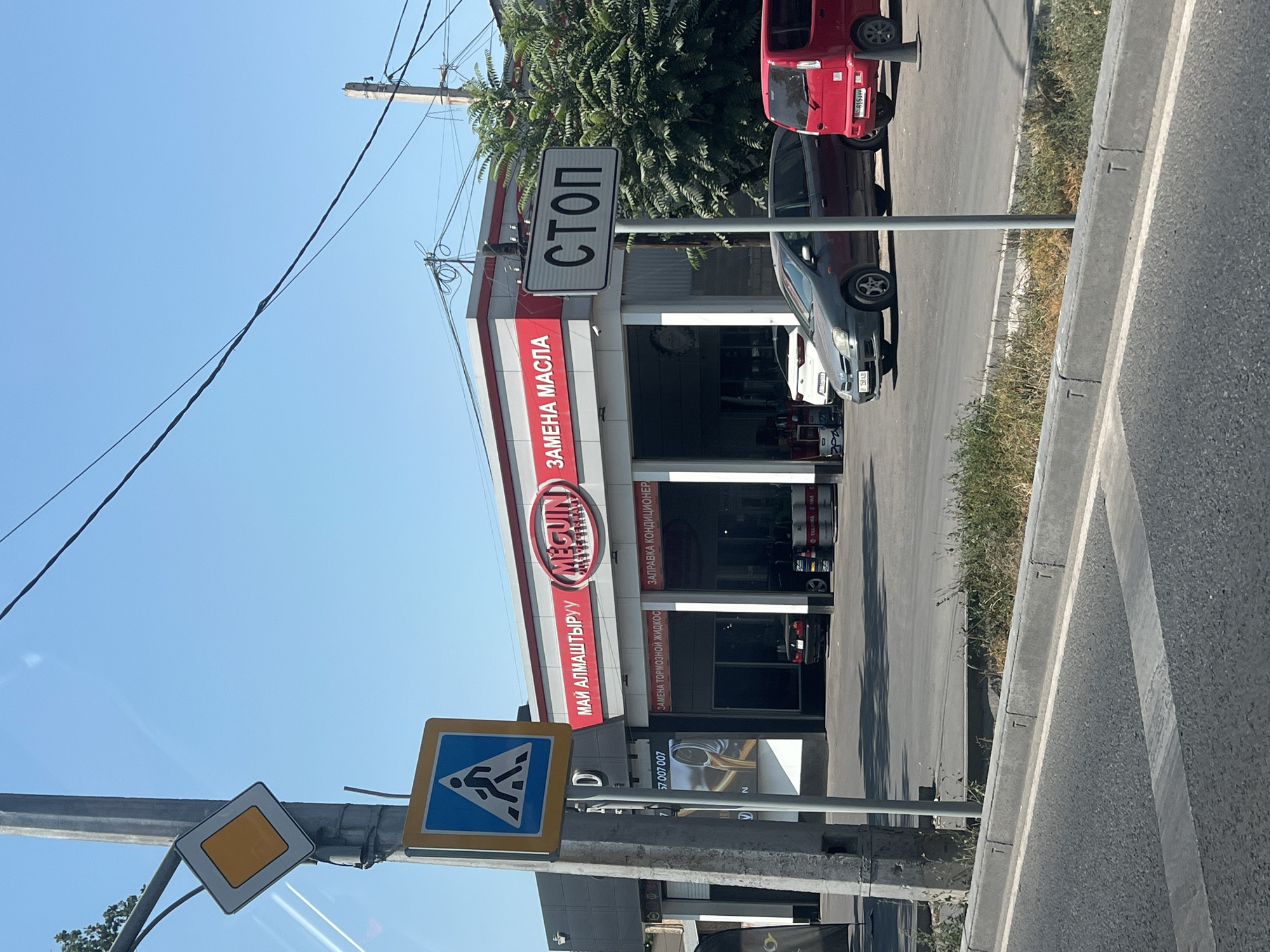 The image shows a street scene in Bishkek, Kyrgyzstan, in front of a service center. A prominent sign with the logo "MEGUIN" and the text "ЗАМЕНА МАСЛА" (Oil Change) is visible on the building. Below this, smaller signs indicate "ЗАМЕНА ТОРМОЗНОЙ ЖИДКОС" (Brake Fluid Change) and "ЗАПРАВКА КОНДИЦИОНЕР" (Air Conditioner Refueling). Two cars are parked in the service bay, a white SUV and a black sedan. A red van is parked on the street in front of the service center.
On the left side of the image, a traffic light pole supports a yellow diamond-shaped sign and a blue square sign depicting a pedestrian crossing. Further down the pole, a phone number is displayed: "57 007 007".
A white pole with a rectangular sign displaying the word "СТОП" (STOP) in black letters is positioned on the right side of the road. The street is paved, with a concrete curb and a strip of overgrown grass and weeds. Power lines are visible against a clear blue sky. The overall scene depicts a typical urban environment with a focus on automotive services.