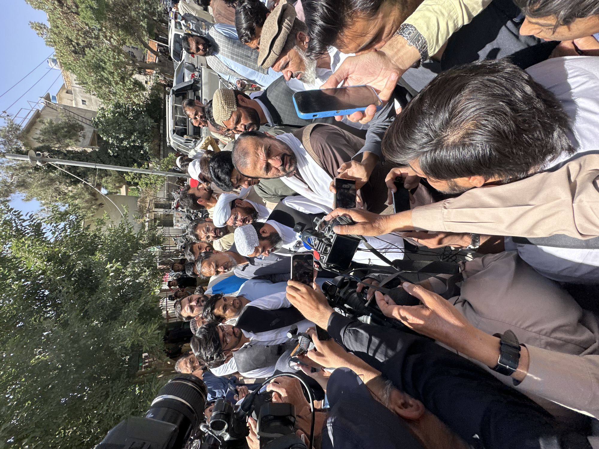 A large group of men has gathered in an outdoor area, presumably in Quetta City Tehsil, Pakistan, judging by the ethnicities of the people in the photo. The group consists of men of varying ages. Many of the men are holding smartphones and cameras, suggesting they are recording or photographing a specific individual or event. In the center of the crowd is a man wearing a white shawl or scarf. The overall atmosphere appears to be intense and potentially chaotic due to the density of the crowd and the activity of the photographers. The location appears to be a street, near trees and buildings.