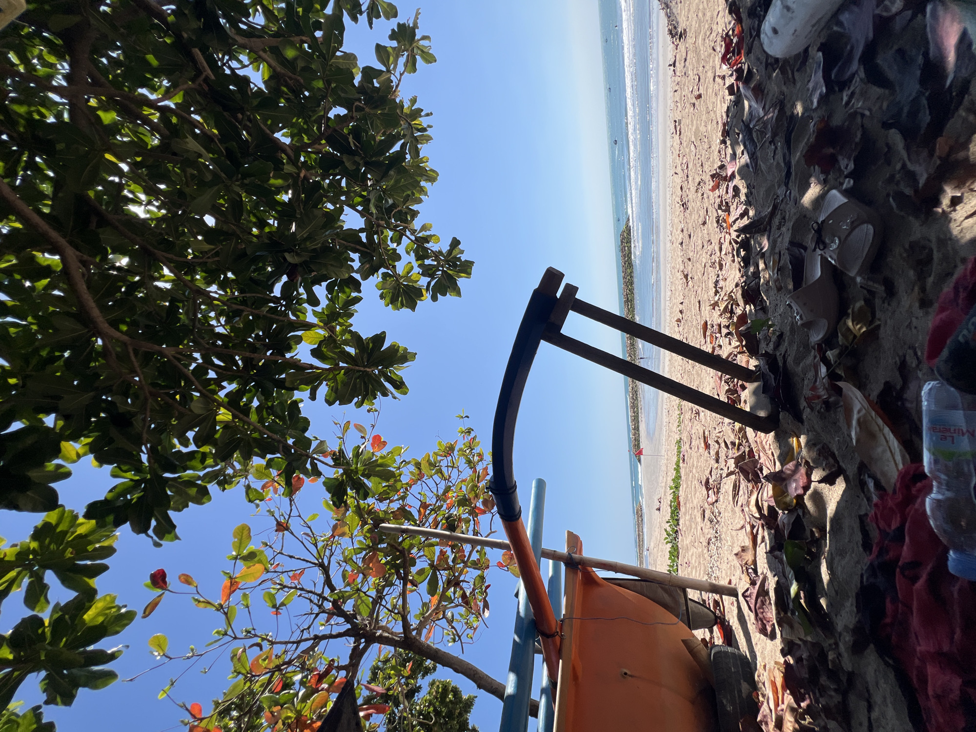 The image depicts a low-angle view of a beach in Kuta, Indonesia, on a clear, sunny day. The foreground is dominated by sand strewn with fallen leaves and a few scattered personal items, including a red cloth, a plastic water bottle, and a pair of flip-flops. Partially visible in the left foreground is the orange hull of a small boat resting on the sand, supported by a wheel and a wooden structure.
In the midground, the sandy beach stretches towards the ocean, with gentle waves lapping at the shore. A line of rocks forms a breakwater or barrier in the sea, and a small red flag is visible on the beach. The water is a clear blue, and several small boats are dotted on the horizon.
The background is a vast expanse of clear blue sky. Overhanging the scene from the top and right is the lush green foliage of a tree, with some leaves exhibiting autumnal colors of red and orange. The overall impression is one of a serene, natural beach setting, likely a quiet spot away from the main tourist areas of Kuta.