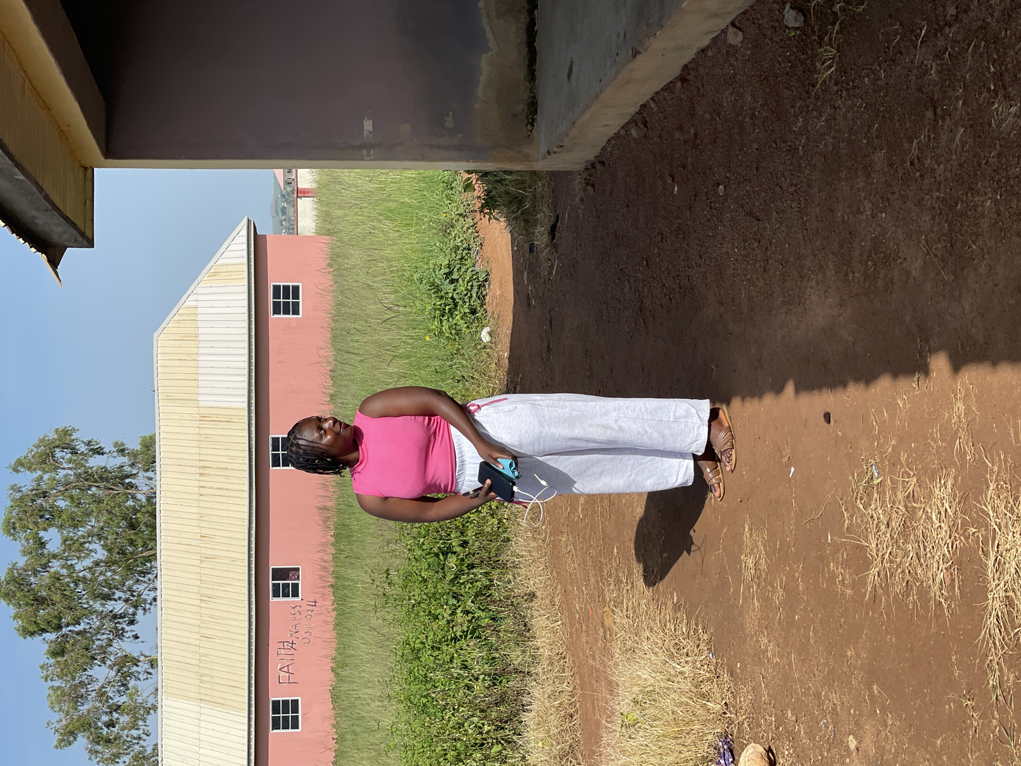 A woman stands in front of a pink building in Durumi, Nigeria. The woman is wearing a pink tank top and grey pants. She is holding a black phone in her right hand and a blue charging cable in her left hand. The building behind her is a single-story structure with a white roof and windows. The area is overgrown with grass and some trees. The ground is covered with dirt and some scattered debris. The sky is clear and blue. The scene is lit by natural sunlight.