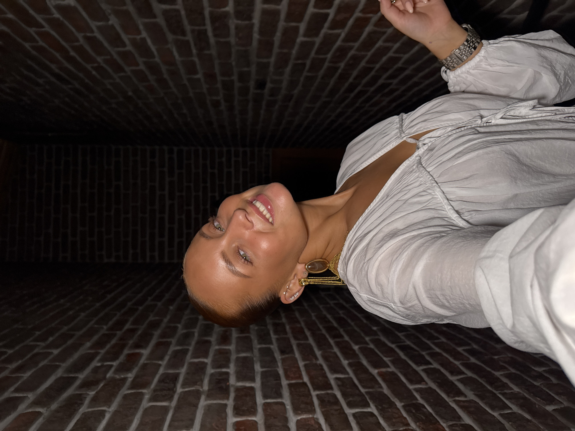 A woman with red hair pulled back and green eyes smiles at the camera. She is wearing a white, open-necked blouse with voluminous sleeves and large, dangling gold earrings. Her left hand, adorned with a silver watch, rests near the camera. She is positioned in what appears to be a narrow hallway with exposed brick walls. The scene suggests an indoor urban environment.