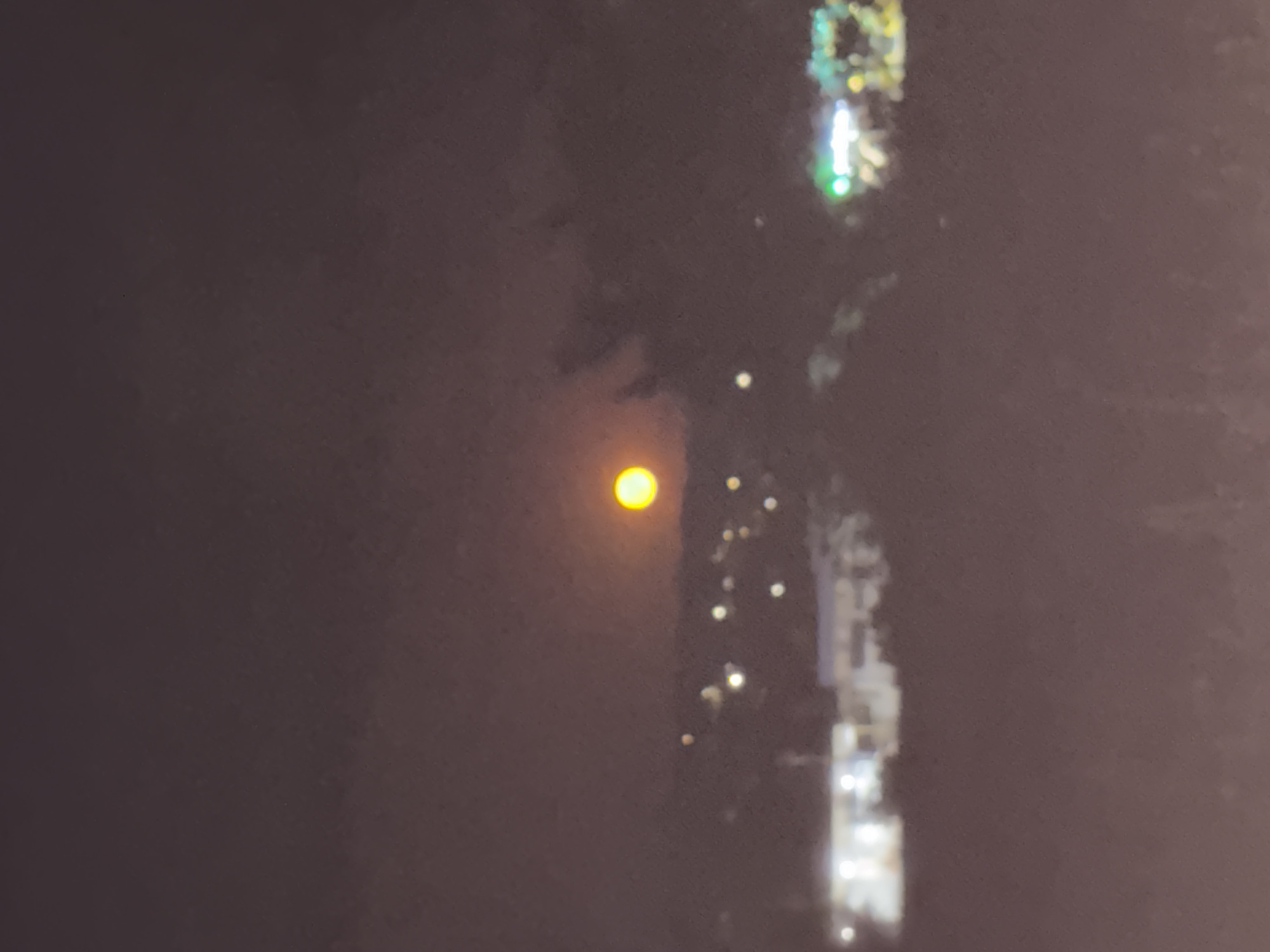 The image captures a nighttime scene in Ceadîr-Lunga, Moldova, with a prominent, hazy orange-yellow moon illuminating the sky. Below the moon, a silhouette of hills or a distant town is visible. Scattered lights, appearing as small, blurred orbs, dot the landscape, suggesting the presence of buildings or streetlights. Some of these lights are white, while others have a greenish hue. The overall atmosphere is dark and somewhat obscured by atmospheric haze or clouds, giving the image a soft, impressionistic quality. No people or specific structures are clearly identifiable due to the low resolution and darkness.