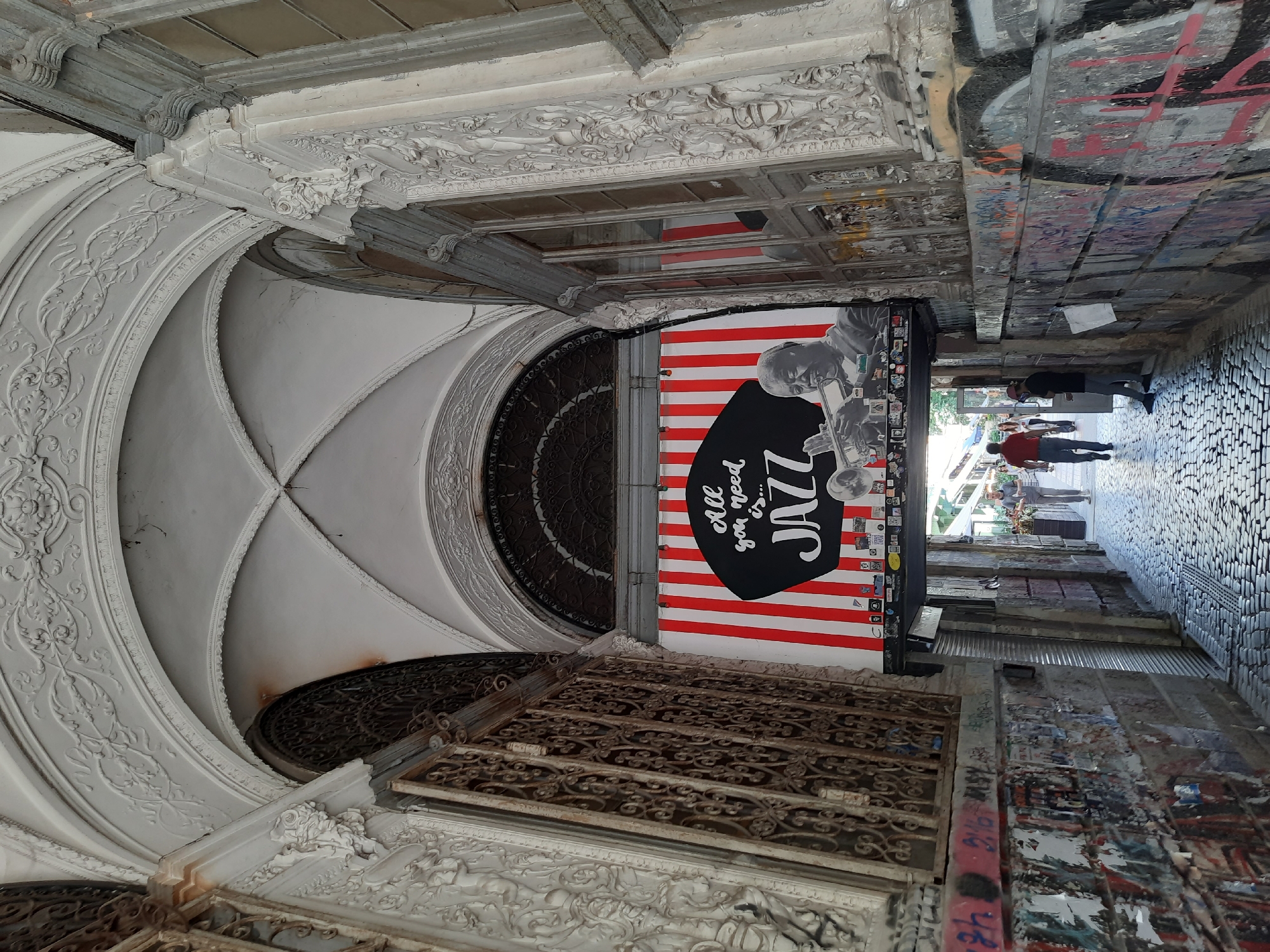 An image shows a view down an arched cobblestone alleyway in Lviv, Ukraine. On the right wall of the alley, a large banner with red and white vertical stripes features a black circular graphic with white text that reads "All you need is... JAZZ". Below the text is a black and white image of a man playing a trumpet. The walls of the alley are covered in graffiti and posters. People are walking down the alley in the distance. The architecture of the alleyway features ornate white plasterwork under the arches.