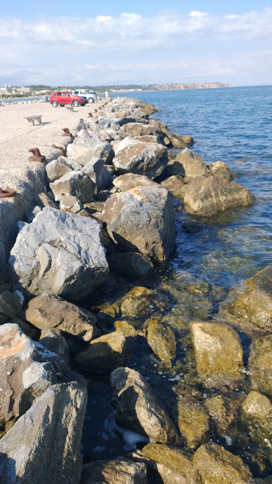 A paved walkway runs parallel to a rocky shore on the coast of Neoi Epivates, Greece. On the walkway, a red SUV and a white SUV are parked. A small, wooden bench is situated on the left side of the walkway. The sea, with its clear blue water, laps against the rocks. The sky above is a bright blue with scattered white clouds. The overall scene depicts a sunny day at a coastal location.