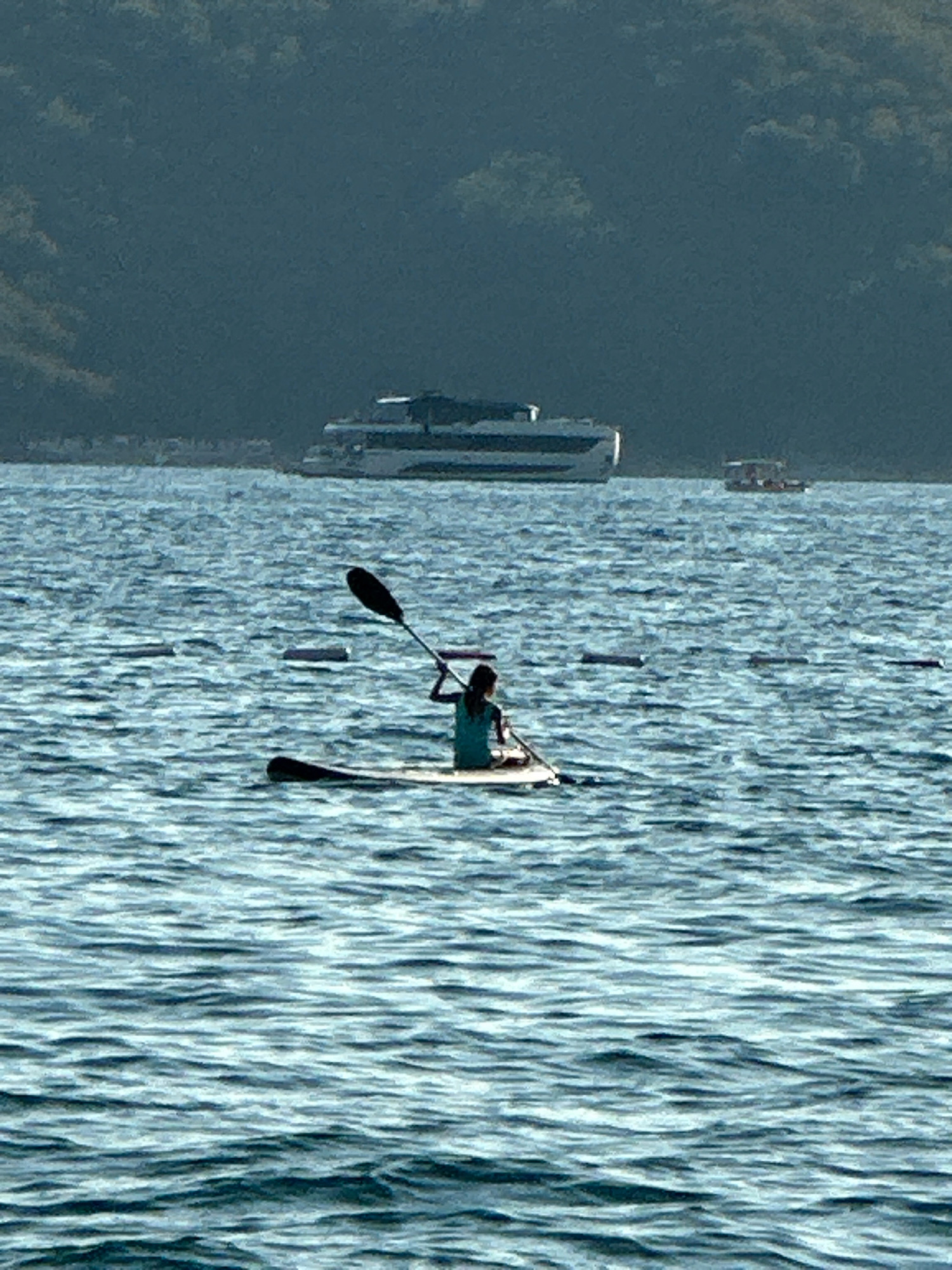 A person is paddleboarding on a body of water. The person is wearing a teal shirt and is holding a paddle. The paddleboard is white. In the background, there are two boats on the water and a forested hillside. The body of water is choppy. The image appears to have been taken on a sunny day. It is not possible to determine the specific location within the city of Boreti, Montenegro from the image alone.