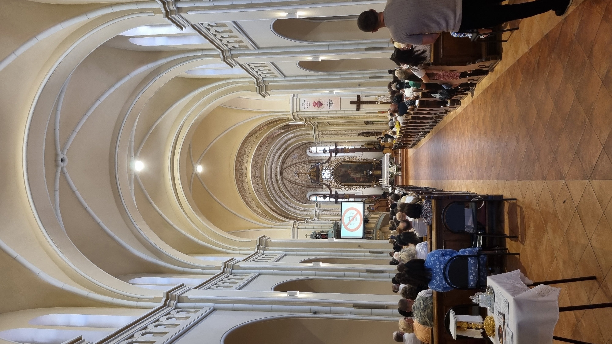 The image shows the interior of a church, likely a cathedral, with attendees seated in pews. The architecture features high, vaulted ceilings with arched supports and ornate detailing. A large painting of a religious scene is visible above the altar. A screen is displaying a "no cell phone" symbol. The church appears to be located in Mukachevo, Ukraine, based on the prompt. The lighting suggests it's daytime. The attendees are dressed casually. A man in the foreground is facing away from the camera.