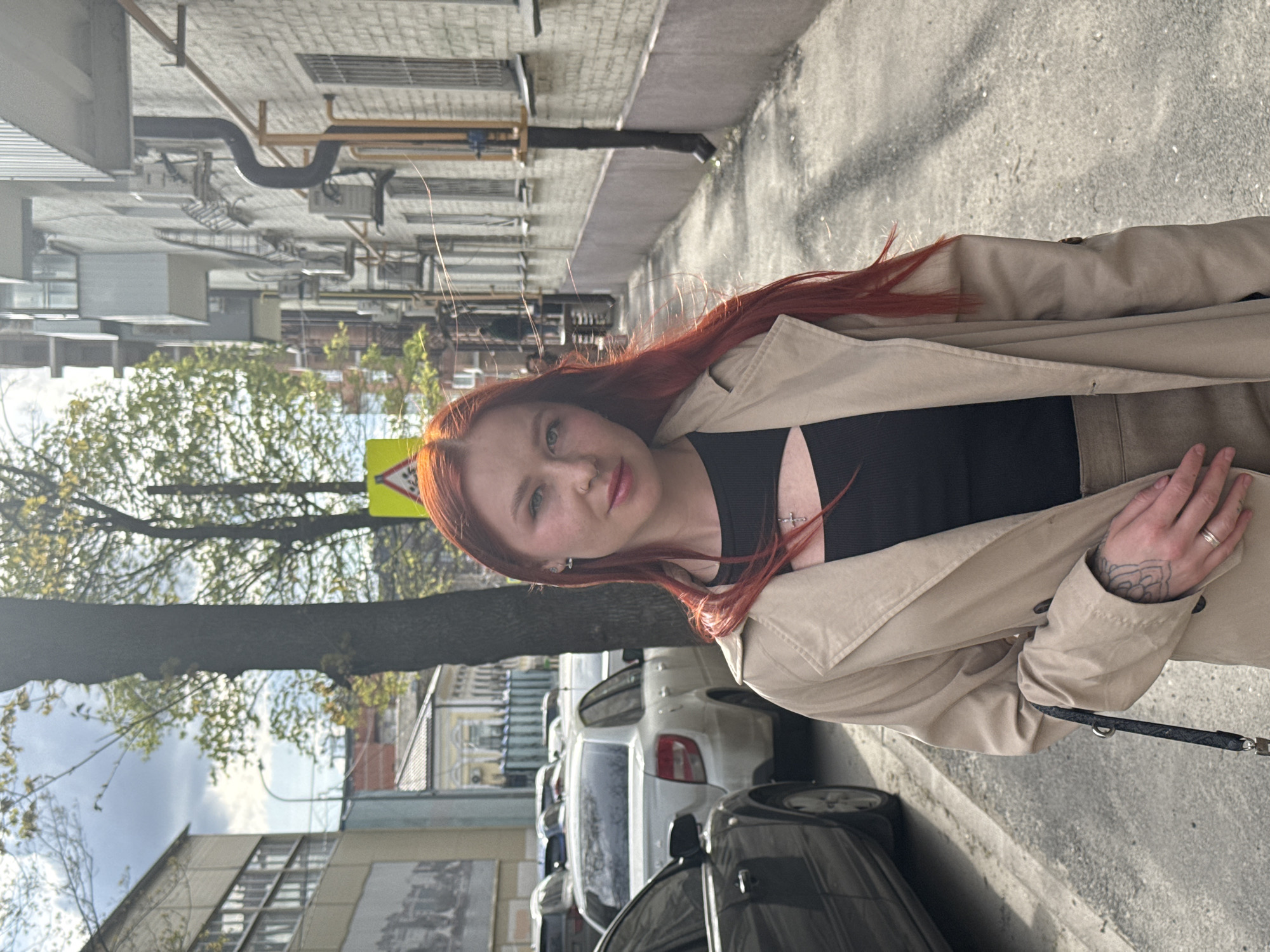 A young woman with red hair, wearing a black top and a beige trench coat, is standing on a sidewalk in Rostov-on-Don, Russia. She is facing the camera with a slight smile. A leash is visible in her hand. Behind her, a row of parked cars lines the street. Buildings and trees are visible in the background. The presence of a road sign typical for Russia suggests the location is likely Rostov-on-Don. The day appears sunny.