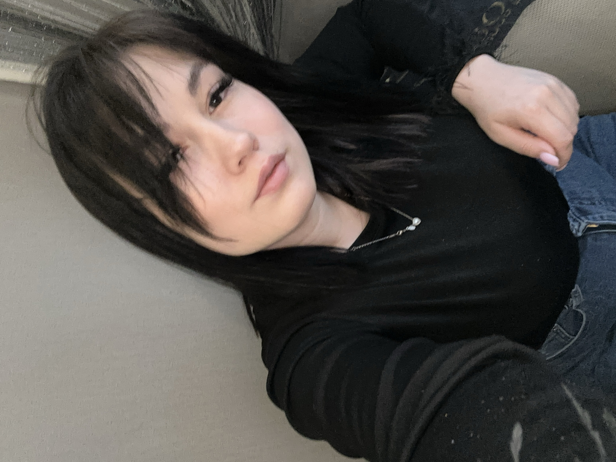 A young woman with dark hair is lying down, possibly on a couch or bed. She is wearing a black top and blue jeans. A necklace is visible around her neck. The background is a light, neutral color. There is no evidence to suggest this image was taken in Chaikovsky, Russia, or any specific location. The scene appears to be indoors.