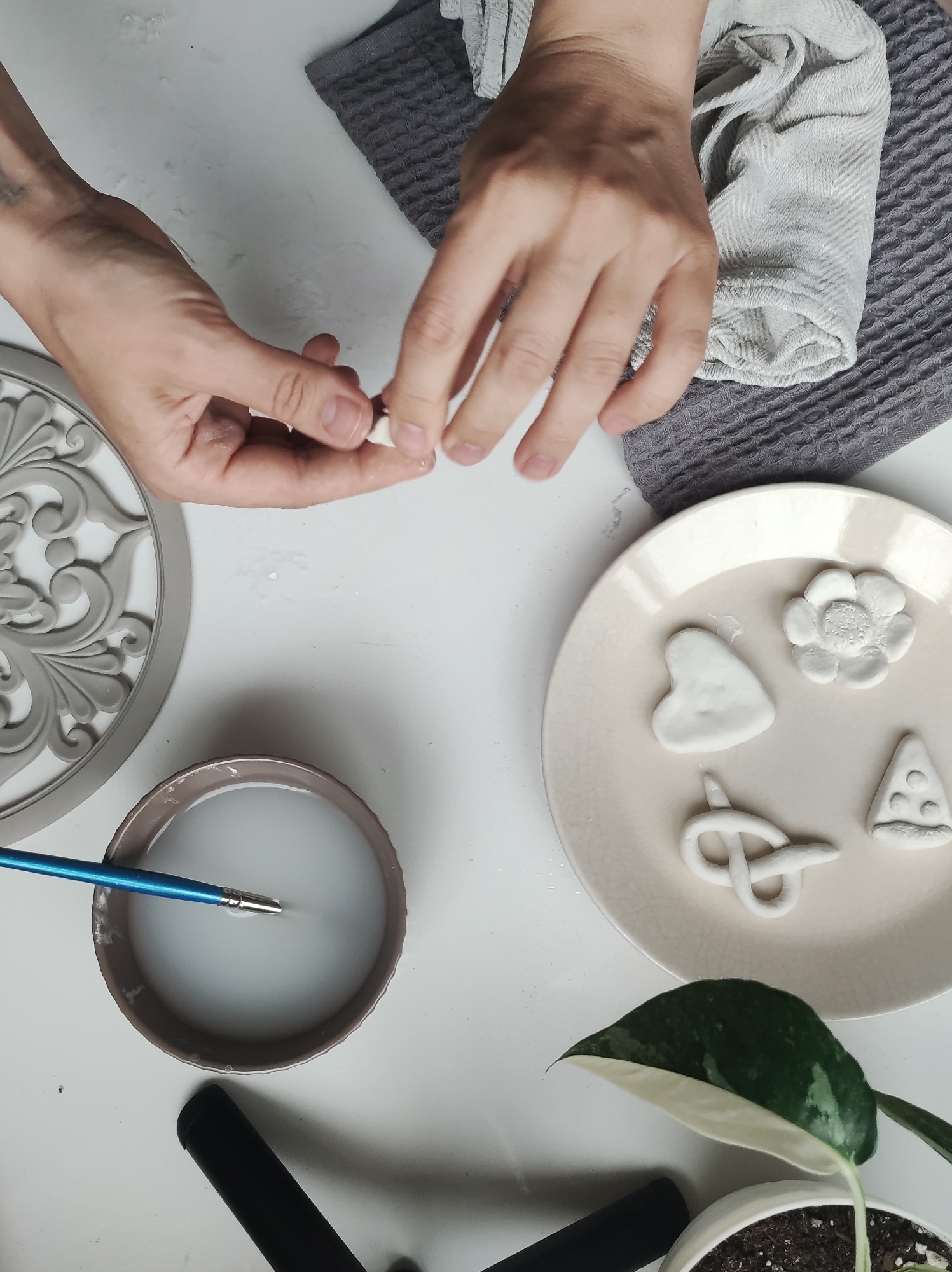 An overhead shot shows a person crafting clay ornaments. The person's hands are visible, working with a small piece of white clay. Various handmade clay ornaments including a heart, flower, pretzel, and pizza slice sit on a cream-colored plate. A bowl of water with a blue paintbrush inside rests nearby. A gray patterned placemat and towel are placed on the white surface alongside a decorative gray resin frame. A plant in a white pot sits in the corner of the frame. It is unknown if this photo was taken in Chaikovsky, Russia.