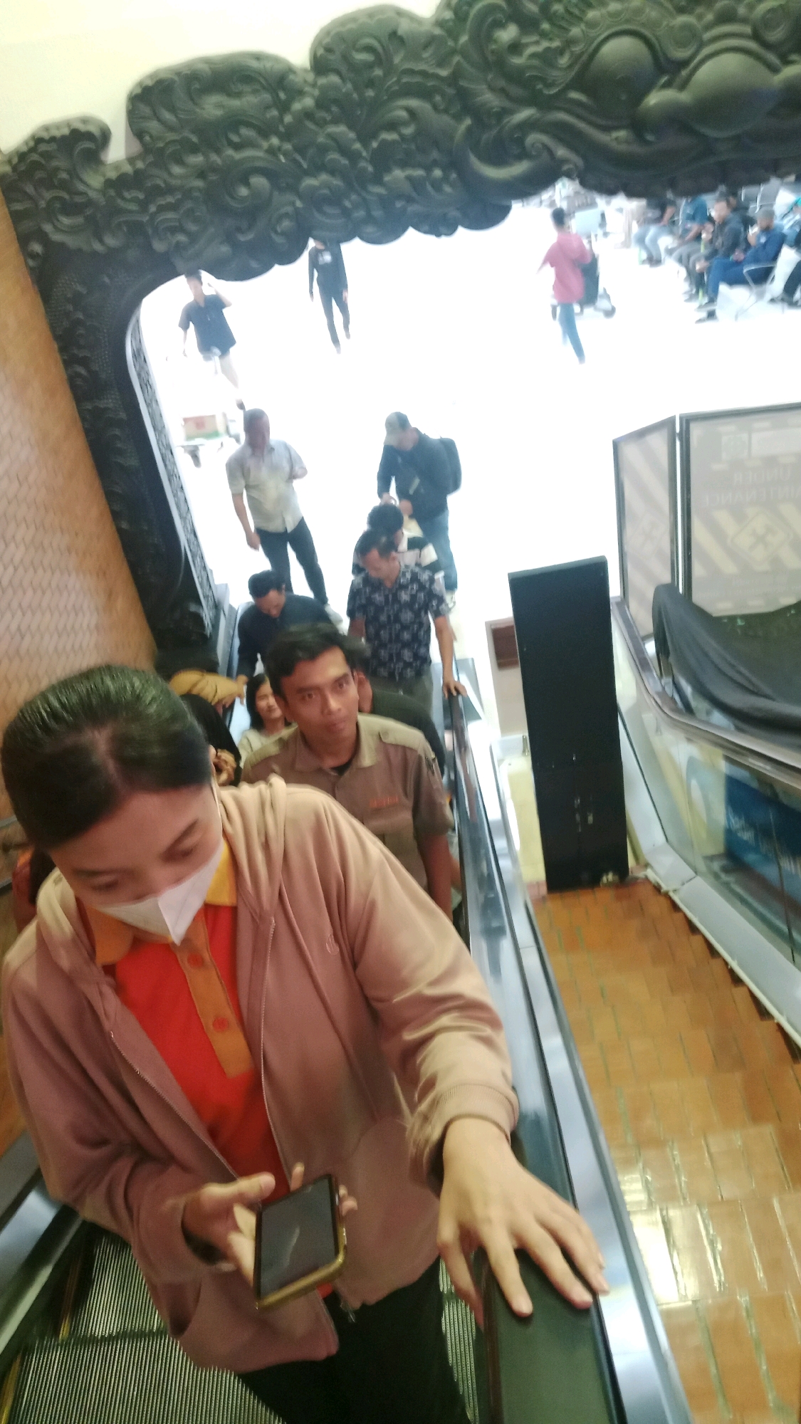 The image captures a group of people on an escalator. In the foreground, a woman wearing a pink hooded jacket over an orange shirt and a white mask is holding a smartphone. Behind her, several other individuals are ascending the escalator, including a man in a beige uniform and a man in a patterned shirt. In the background, more people are visible, some walking and others seated. The scene is framed by an ornate, dark decorative archway above. The floor appears to be tiled. Based on the style of the archway and the general setting, this location is likely in Tangerang, Indonesia, possibly within a shopping mall or a similar public venue that features elaborate architectural elements.
