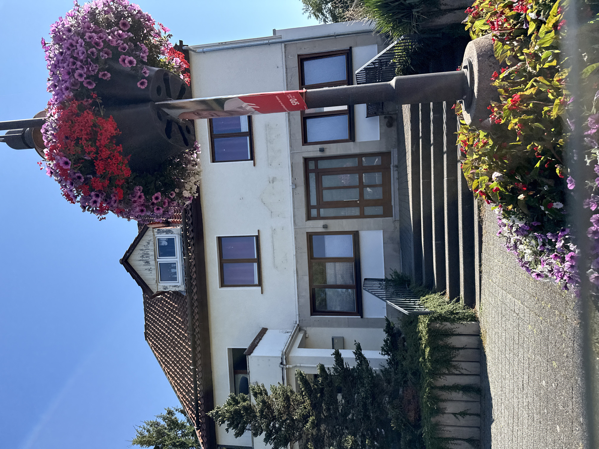 The image depicts the exterior of a white building with a red tiled roof, situated in the city of Bad Sachsa, Germany. The building features multiple windows with brown frames and some appear to have blinds. A set of stairs with a black metal railing leads up to the entrance, which has a wooden double door. To the left of the entrance, a large evergreen tree stands next to a white balcony. In the foreground, a lamppost is adorned with a large hanging basket overflowing with colorful flowers, including pink, red, and purple petunias. Smaller flower beds with red and purple flowers surround the base of the lamppost. A red banner with a picture and text in German, including "SPD", is attached to the lamppost. The environment suggests a daytime scene with a clear blue sky.