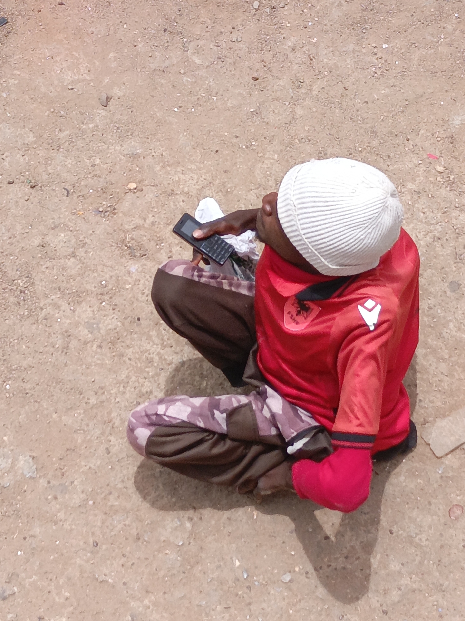 In a medium shot taken from above, a young man is seated cross-legged on a sandy ground. He is wearing a white ribbed beanie, a red long-sleeved shirt with an Albanian emblem, and brown and camouflage patterned trousers. He is holding a black mobile phone in his left hand and looking upwards. The location is Jos, Nigeria, based on the background and attire. The environment is a sandy or dusty open space.