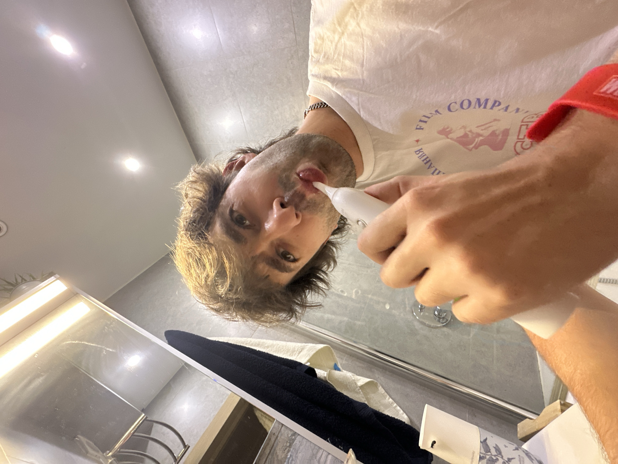 A man with light brown, tousled hair is brushing his teeth with an electric toothbrush in a bathroom. He is wearing a white t-shirt with a circular graphic and text in purple and pink. A silver chain is visible around his neck. His left arm is resting on a white object. To the left, a dark blue towel hangs over a rack, and a white towel with blue stripes is draped over it. A dispenser with a floral pattern is also visible on the counter. The walls are tiled in a light gray color, and the ceiling has overhead lighting. The scene is shot from a low angle, with a reflection of the man and the bathroom fixtures visible in a mirror on the left side of the frame. The overall impression is of a candid, everyday moment captured in a private setting.