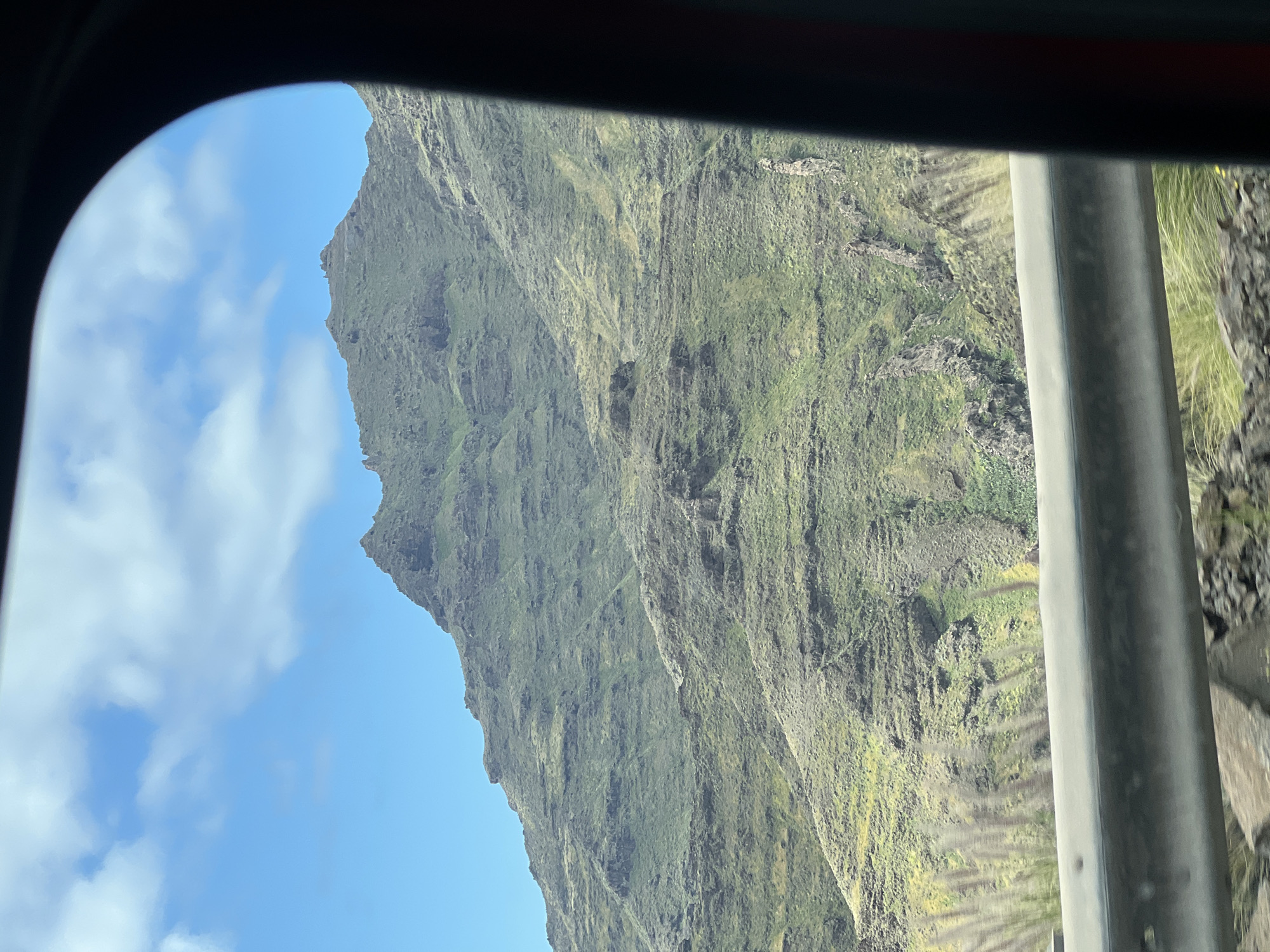 View through a window, possibly of a vehicle, showing a steep, green, mountainous landscape, likely a cliff face. The sky with scattered clouds is visible in the left part of the frame. The mountains are covered with vegetation and appear rugged, with visible rock formations and ravines. A metal pole or structure is present to the right of the frame. This is likely in the municipality of Mogán, Gran Canaria, Spain, as the island is known for its dramatic cliffs and mountainous terrain.