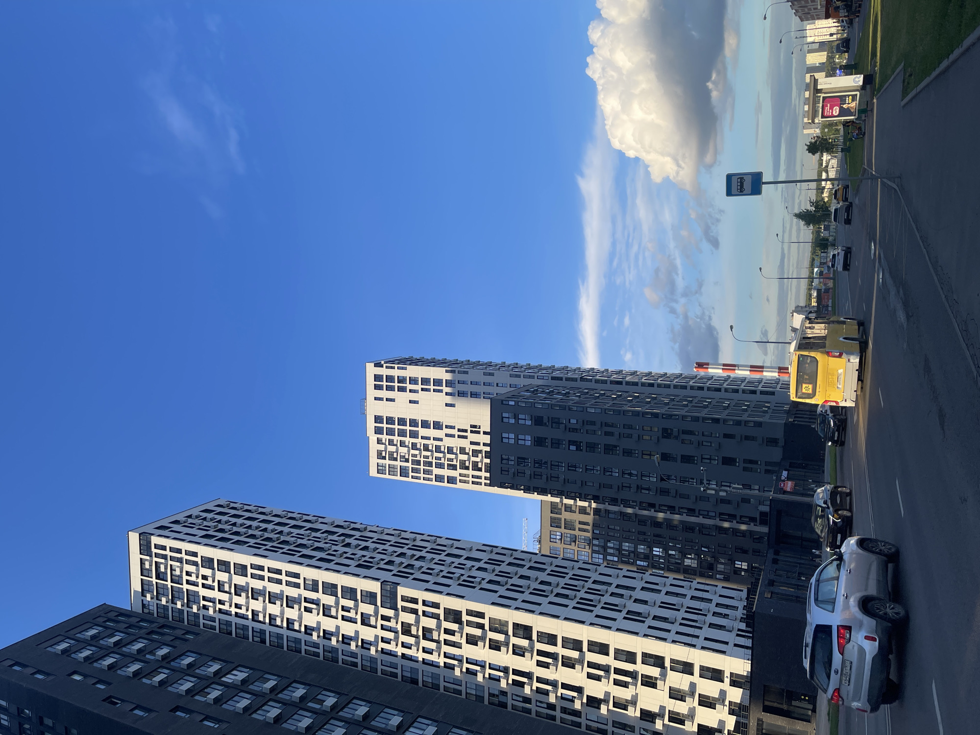 This media file captures an urban scene in Moscow, Russia, under a bright blue sky with scattered clouds. The foreground features modern, tall residential buildings with a distinctive grid-like facade of windows. One building is predominantly dark gray, while the other is white with dark window frames. Sunlight casts strong shadows on the buildings, indicating it is likely daytime, possibly late afternoon given the angle of the light.
To the right of the buildings, a paved road runs parallel. Several cars are visible on the road, including a silver SUV and a yellow bus. The yellow bus has a sign indicating a bus stop. Streetlights line the road, and a bus stop sign, a blue rectangle with a white bus icon, is prominently displayed on a pole. Beyond the road, there is a grassy area and more buildings in the distance, along with billboards. The scene suggests a typical day in a residential area of a large city, with people commuting or going about their daily activities. No specific emotions are conveyed, but the overall impression is one of a bustling, modern urban environment. No legible text is visible on the buildings or billboards.