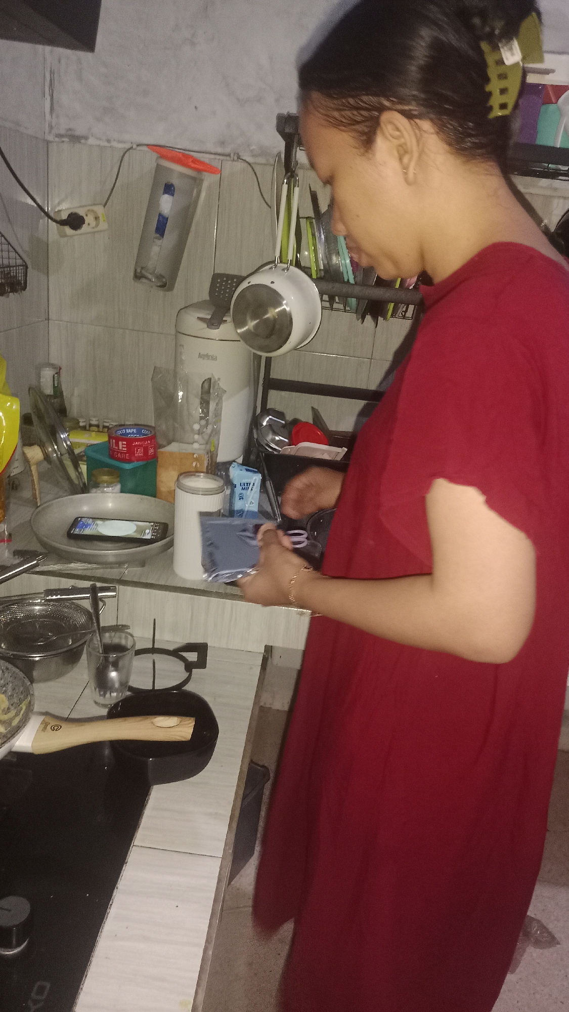 A woman wearing a red shirt stands in a kitchen, holding a dark object, possibly a phone. The kitchen has tiled walls and a countertop with various items, including a rice cooker, a glass, a dish, a spatula, and a phone that is face down on a plate. A power outlet is visible on the wall. The scene appears to be set in a residential kitchen.