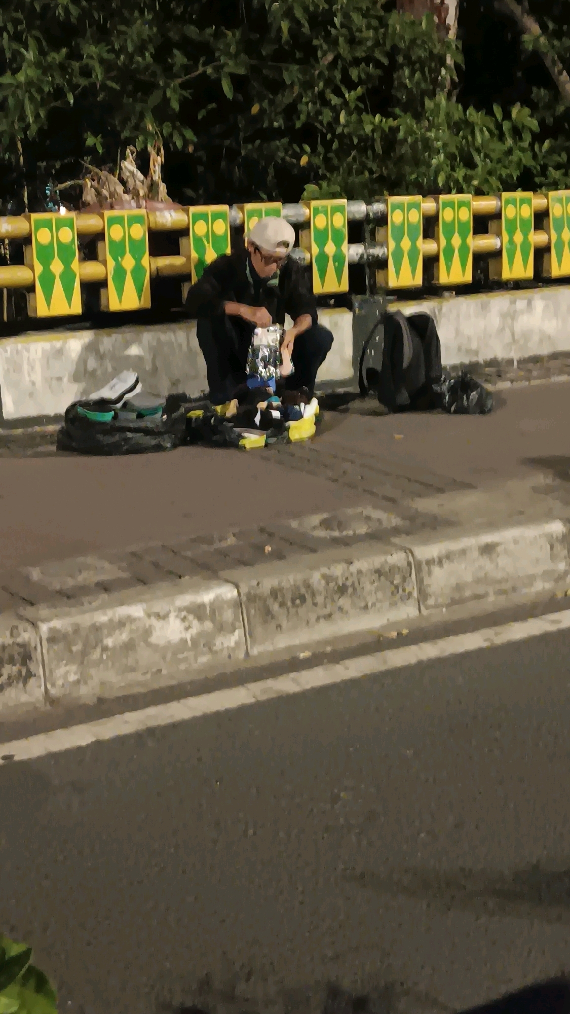 A person wearing a dark jacket, dark pants, and a light-colored baseball cap kneels on a paved sidewalk next to a street at night. The person is looking down at a collection of items laid out on black plastic sheeting. Among the items are several pairs of shoes, including white athletic shoes and dark-colored shoes, as well as some brightly colored flip-flops or sandals. A black backpack is visible behind the kneeling person. The sidewalk is bordered by a concrete curb and a street. In the background, a yellow and green railing with a decorative pattern separates the sidewalk from an area with trees and foliage. The scene suggests the person may be selling or organizing items on the sidewalk. The railing design is typical of public infrastructure in Central Jakarta, Indonesia.