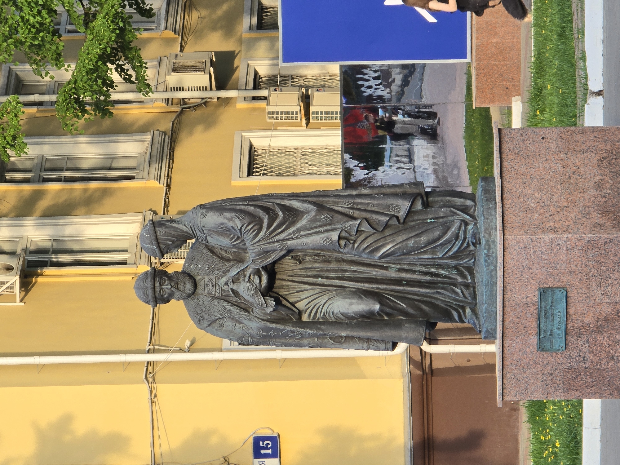 The image shows a statue of two figures in Khimki, Russia. The statue is made of a dark metal and depicts two figures in robes. The statue is located on a reddish-brown stone base with a plaque. Behind the statue is a yellow building with white-framed windows and several air conditioning units. A blue sign is partially visible above the statue. A patch of green grass is visible to the right of the statue's base. A street sign with the number "15" is affixed to the building next to the statue.