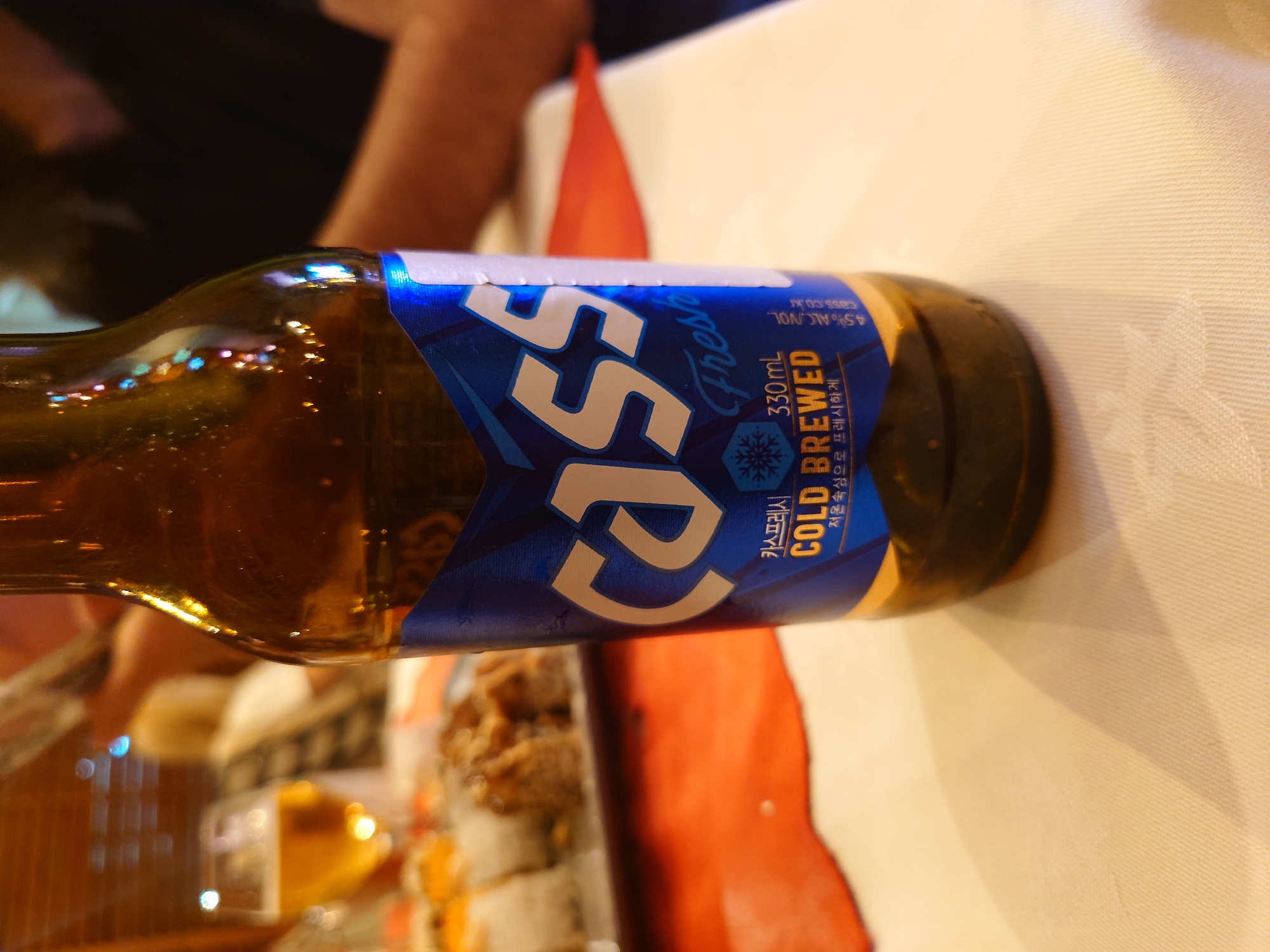 A bottle of Cass Fresh beer is prominently featured, lying horizontally on a white tablecloth, likely in an indoor setting such as a restaurant or bar in Kozloduy, Bulgaria. The brown glass bottle has a distinctive blue label with white and yellow text. Large white letters spell "CASS," accompanied by "Fresh" in a stylized script and "COLD BREWED" in yellow. Other visible text includes "카스프레시," "330mL," "저온숙성으로 프레시하게" (meaning "freshly made with low-temperature aging"), "4.5% ALC./VOL.," and "cass.co.kr."
The bottle's surface reflects multiple artificial lights, suggesting an evening or nighttime ambiance. A vibrant red napkin or mat is partially visible beneath and beside the bottle, adding a splash of color to the otherwise neutral table setting. In the soft, blurred background, hints of food, possibly sushi rolls, and other indistinct table items indicate a meal or social gathering. Parts of human arms or hands are also faintly discernible in the background, further implying the presence of people. The lighting is dim, focusing attention on the illuminated beer bottle.