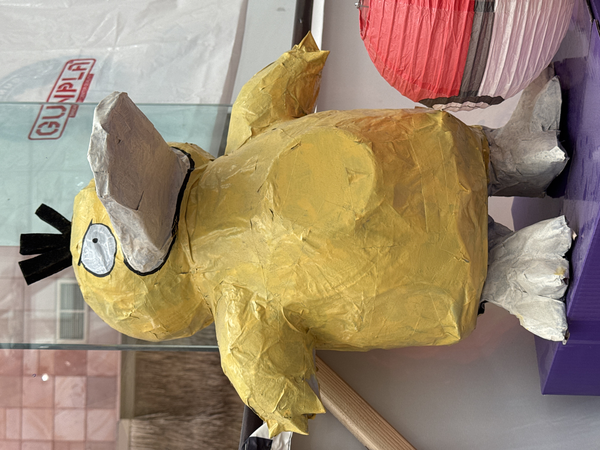 A yellow, paper-mâché figure of the Pokémon Psyduck stands on a purple rectangular platform in El Paso, United States of America. The figure is approximately three feet tall and appears to be a piñata, evidenced by the textured paper covering and the visible seams. Psyduck's beak is made of a light gray, crinkled material, and its eyes are large and white with black pupils. The figure has black, spiky hair on its head. To the right of the Psyduck figure is a red and white spherical object, possibly a lantern or another decorative item. The background consists of a glass display window, through which hints of interior elements like shelves and possibly a retail environment are visible. A red and white sign with the word "GUNPLA" in red lettering is partially visible in the upper right portion of the image, suggesting the location may be a store selling models or toys. The lighting is bright, creating reflections on the glass.