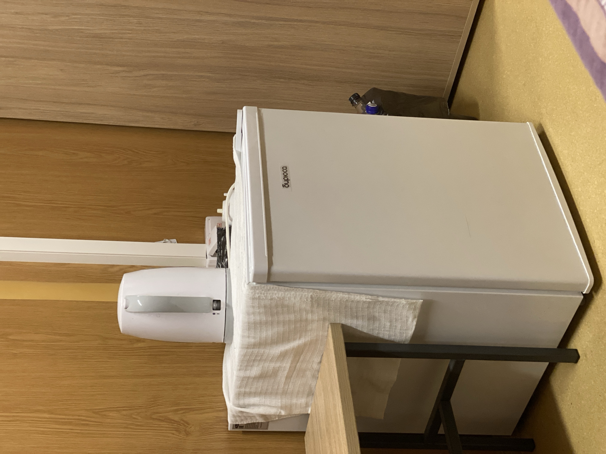The image captures an indoor scene in Koskolovo, Russia, featuring a compact white refrigerator and another white appliance, possibly an air purifier or small air conditioner, resting on a wooden surface. The refrigerator, positioned upright, has "cooling" printed vertically on its door. A light-colored, textured cloth is draped over the wooden surface, extending from underneath the second appliance and partially obscuring the lower side of the fridge.
On top of the refrigerator, a dark-colored plastic bottle with a blue cap lies horizontally. The background consists of natural wood paneling, presenting a warm, earthy tone. Above the lower wooden panel, a lighter wood-grained surface, possibly a cabinet or shelf, is visible. To the right of the appliances, a textured, yellowish wall or cork board is partially visible. In the lower right corner, a black metal frame, likely part of a table or shelving unit, supports the wooden surface. No specific activity or event is taking place; the image shows a static arrangement of items. The setting appears to be a small living space, such as a dorm room or a compact apartment. The time of day, weather, and emotions cannot be determined from the photograph.