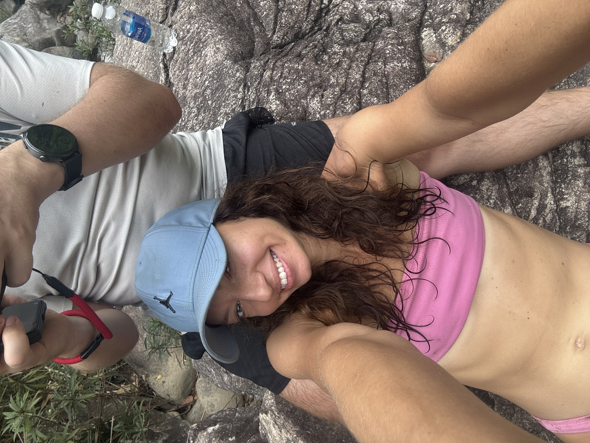 A selfie depicts a woman in a pink top and blue baseball cap lying on rocks, with two people visible beside her. One individual is wearing a white shirt and a black smartwatch, holding a black object with a red strap. A bottle of water sits nearby. The location appears to be outdoors, possibly a rocky area near foliage. While the specific location in Đà Nẵng, Vietnam, cannot be determined from the image, the setting suggests a natural, possibly mountainous area within or near the city.