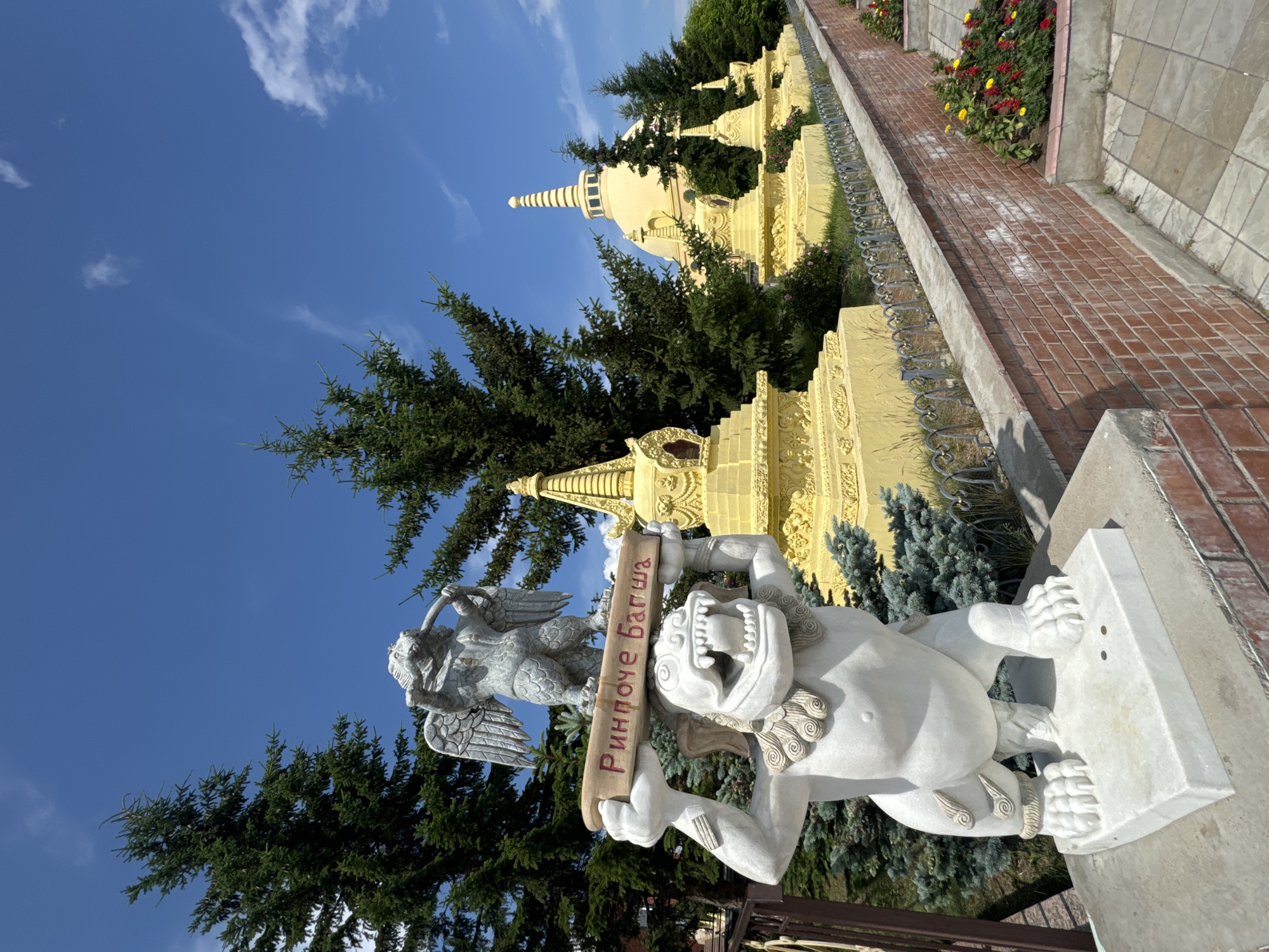 This is a photograph of the Buddhist Temple complex in Ulan-Ude, Russia, under a bright blue sky with scattered white clouds. In the foreground, a white marble statue of a mythical creature with a lion-like body and an eagle's head stands on a pedestal. It is holding a wooden beam across its shoulders, which is inscribed with red Cyrillic letters that read "Ринпоче Багша" (Rinpoche Bagsha). Behind this statue, a series of yellow stupas, traditional Buddhist structures, are arranged in a line, leading towards a larger, golden-domed temple in the background. Evergreen trees are visible throughout the scene, and a low brick wall borders a flower bed with colorful flowers. The overall impression is one of religious and architectural significance within a natural setting.