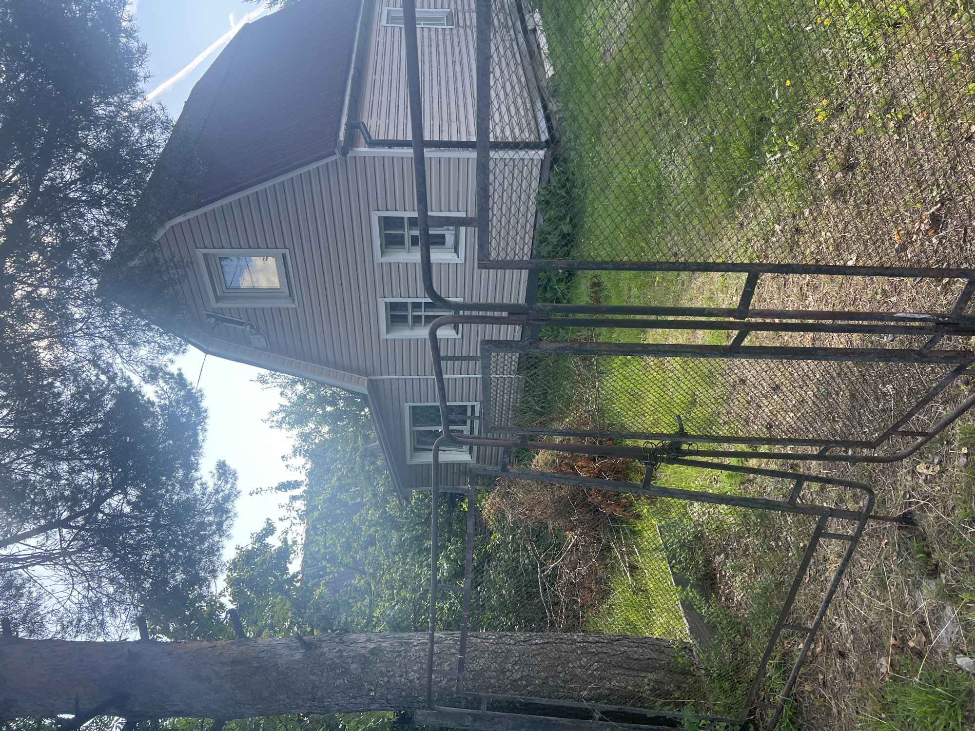 A residential property in Imeni Morozova, Russia, features a two-story house with light beige siding and a dark red metal roof. The house has multiple windows with white trim. In the foreground, a rusty metal gate and chain-link fence partially obstruct the view of the property. A large pine tree stands to the left of the gate, with its trunk visible. The ground is covered with grass and dry leaves. Lush green trees and foliage surround the house, and the sky is blue with scattered clouds. The image appears to have been taken on a sunny day.